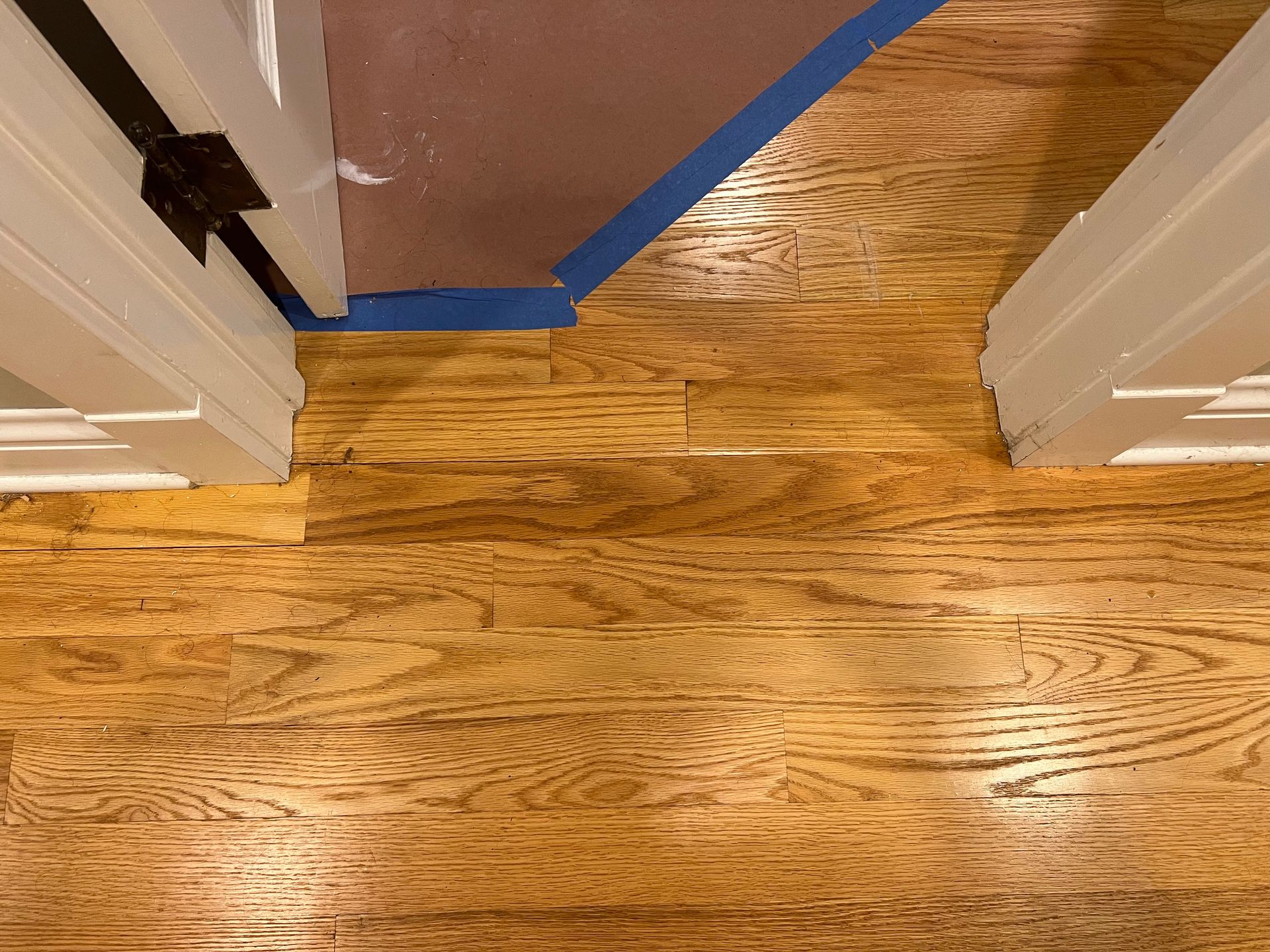 Wooden hallway floor with blue painter’s tape along the edges near a doorway under renovation
