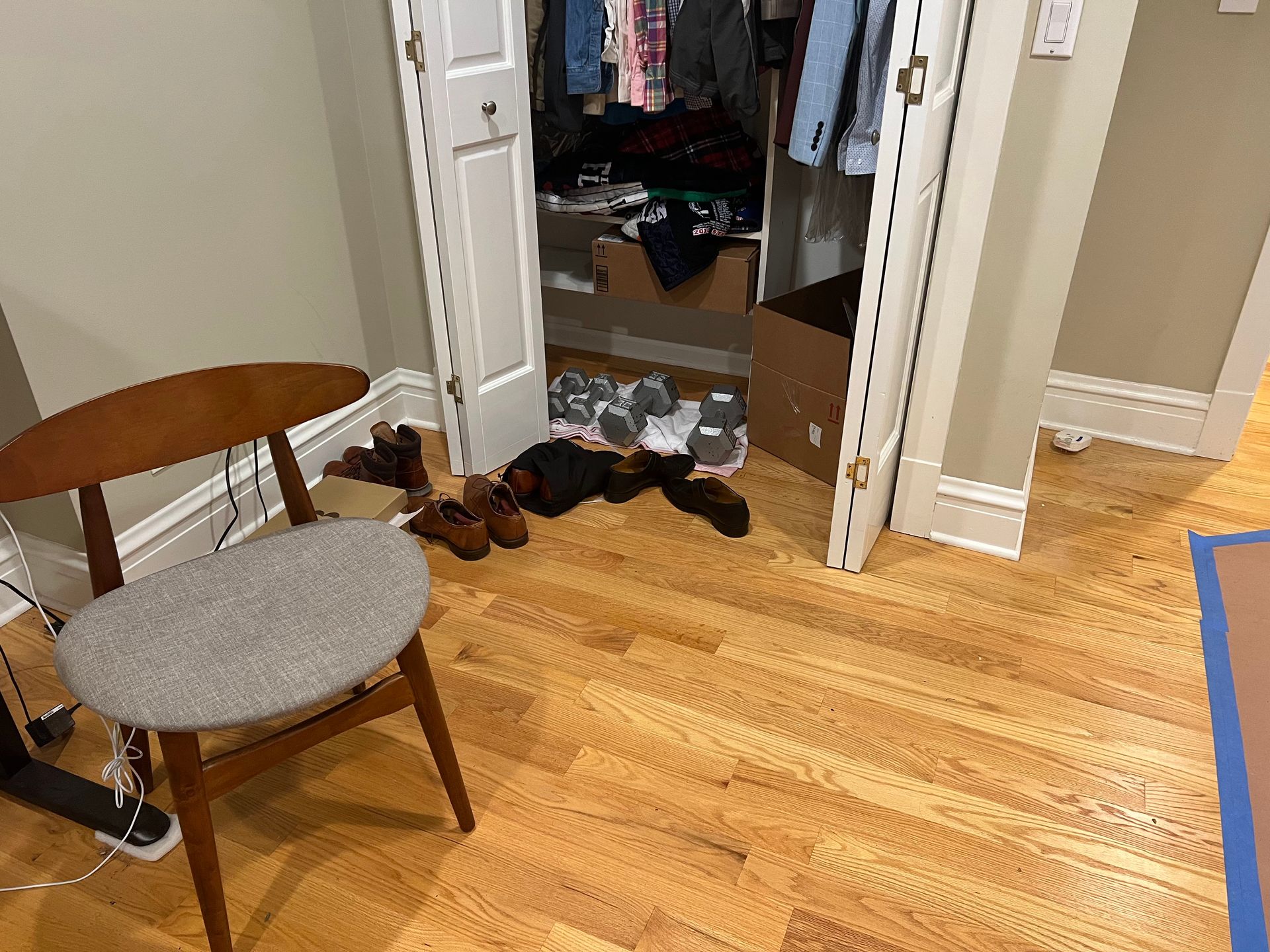 Open closet doorway with shoes on the floor, chair at left, and hardwood floor in a hallway.