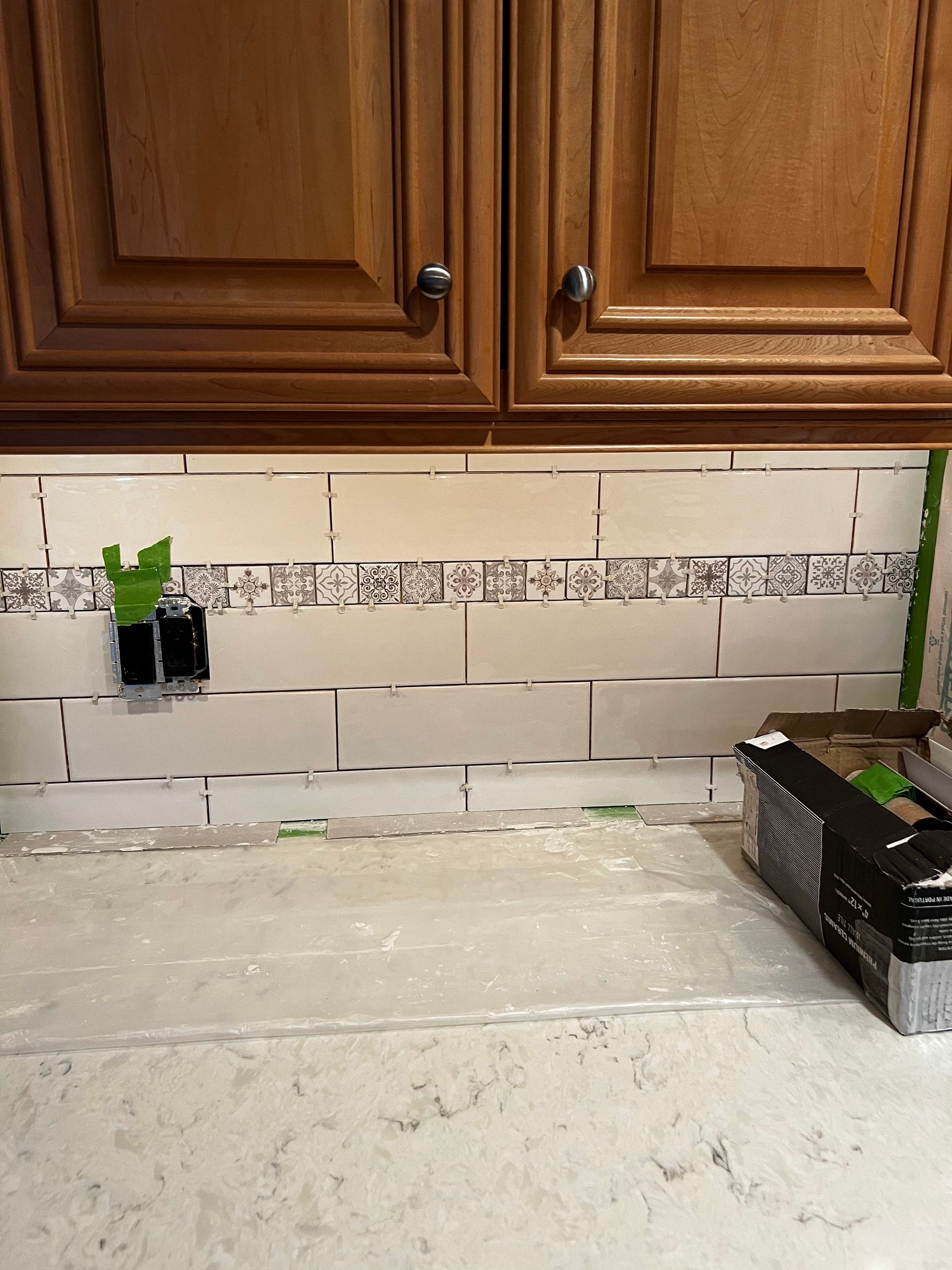 Kitchen countertop with white tile backsplash, wooden cabinets, and a small green wall outlet cover.