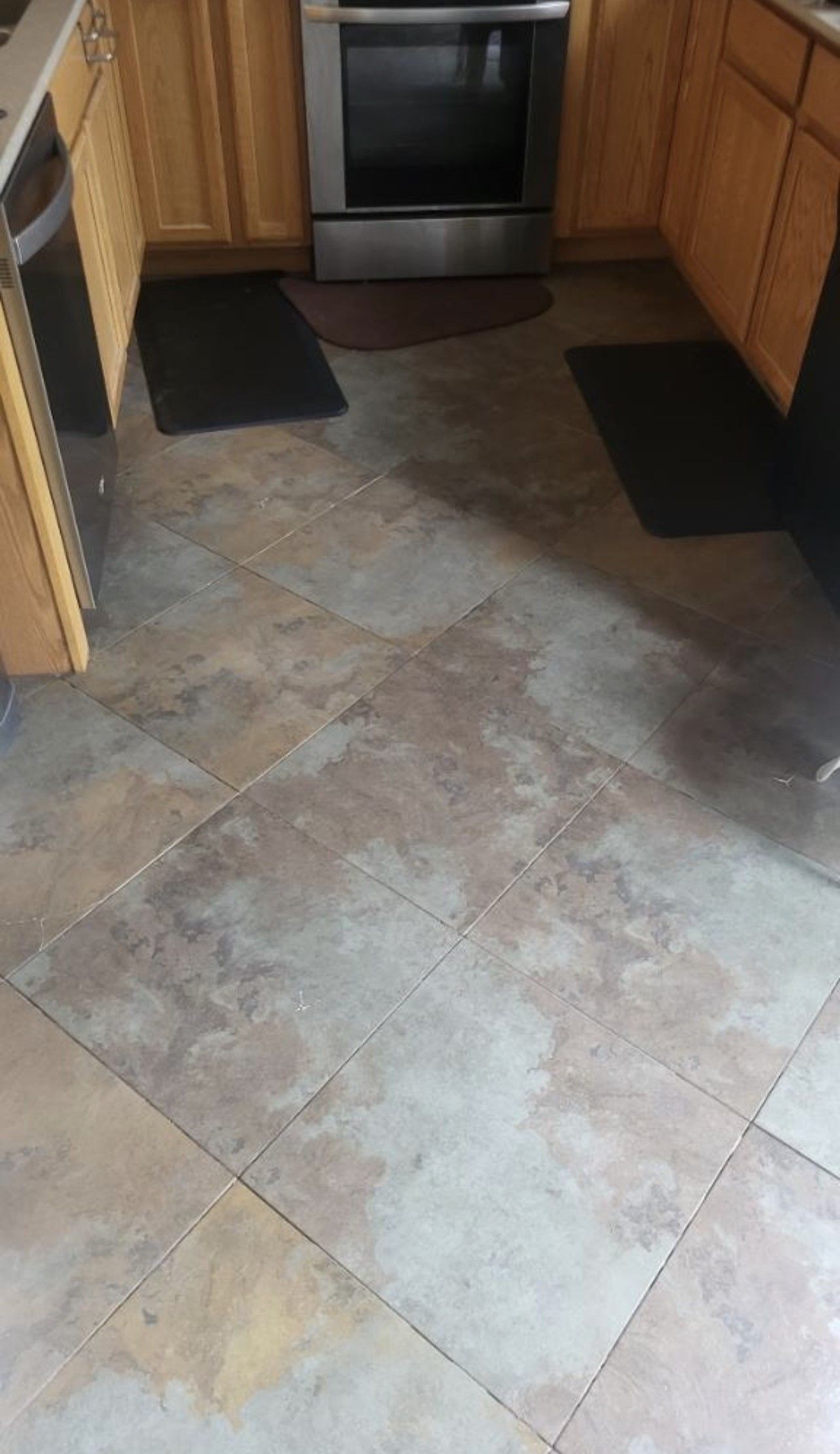 Kitchen floor with large beige-gray tiles, black mats, and wooden cabinets by an oven.