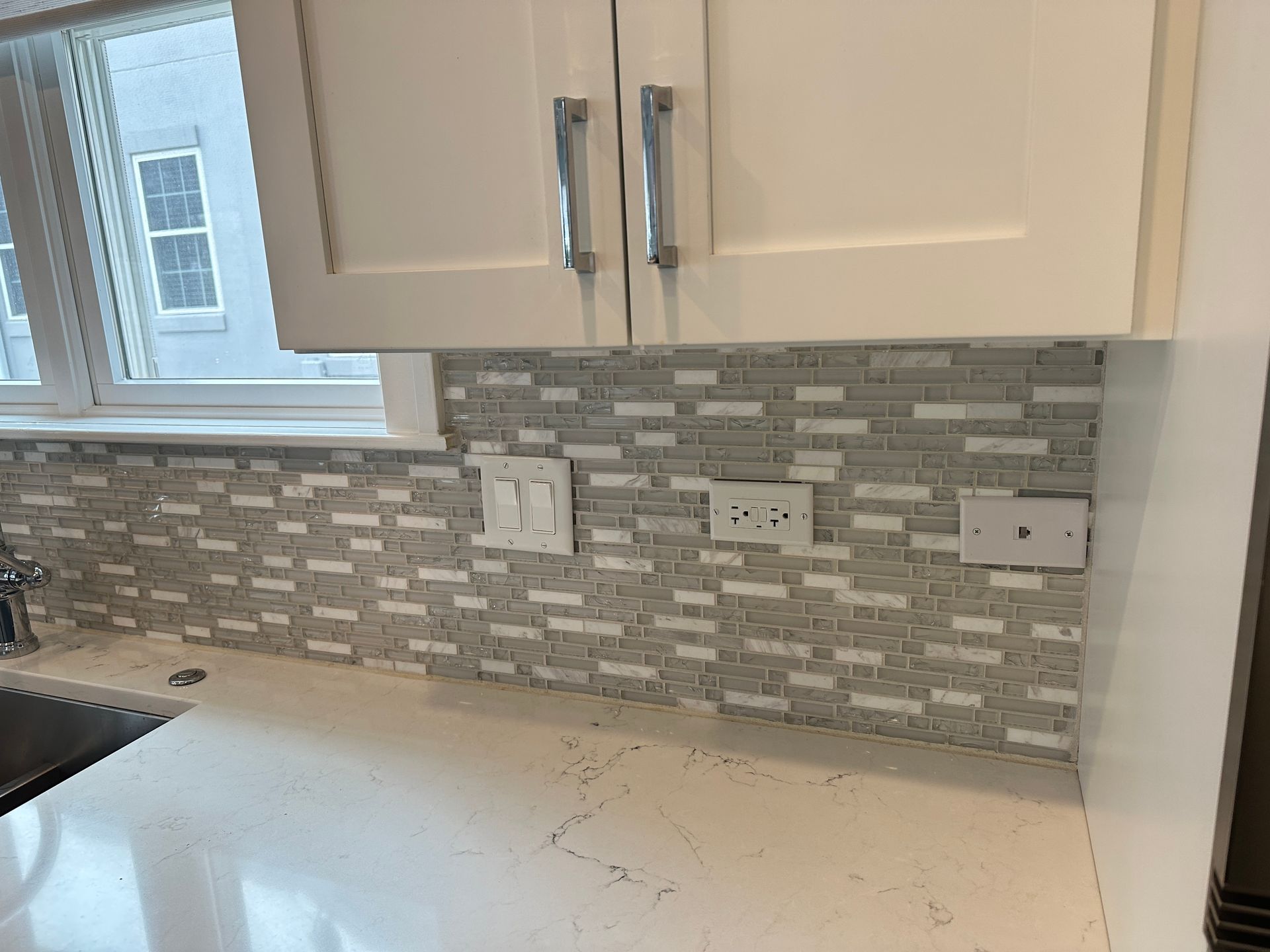 Kitchen counter with light cabinets, mosaic backsplash, window, and outlet cover missing on the right wall.