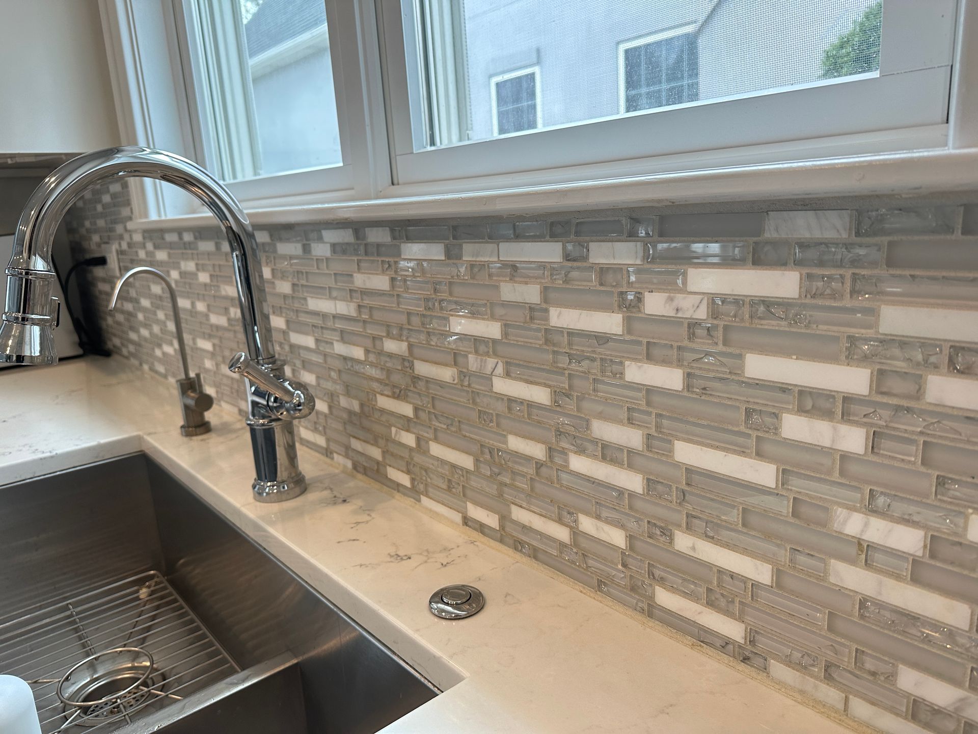 Kitchen sink with chrome faucet, light countertop, and gray tile backsplash under a window.