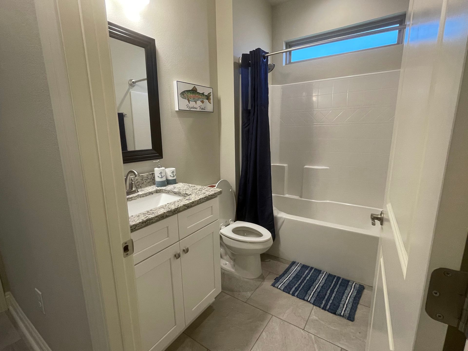 Small bathroom with vanity, mirror, toilet, shower-tub, blue curtain, and striped bath mat.