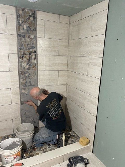 Worker installing wall tiles in a bathroom, kneeling beside buckets and tools.