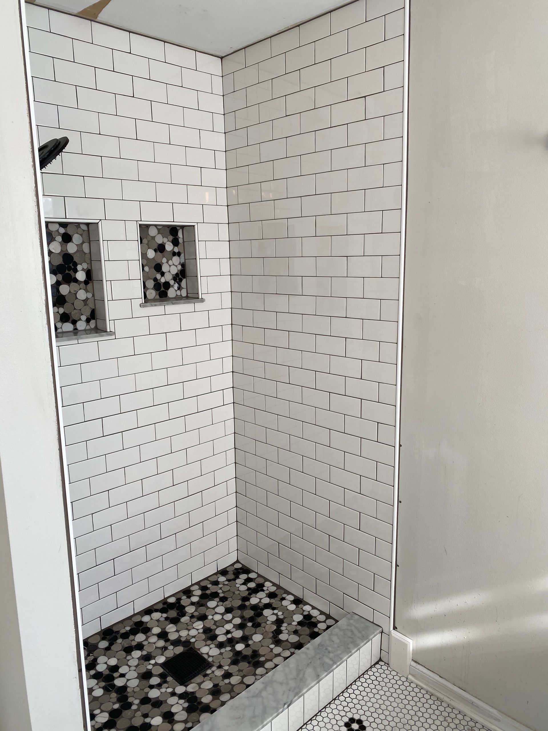 White-tiled shower stall with black mosaic floor and two wall-mounted shower valves.