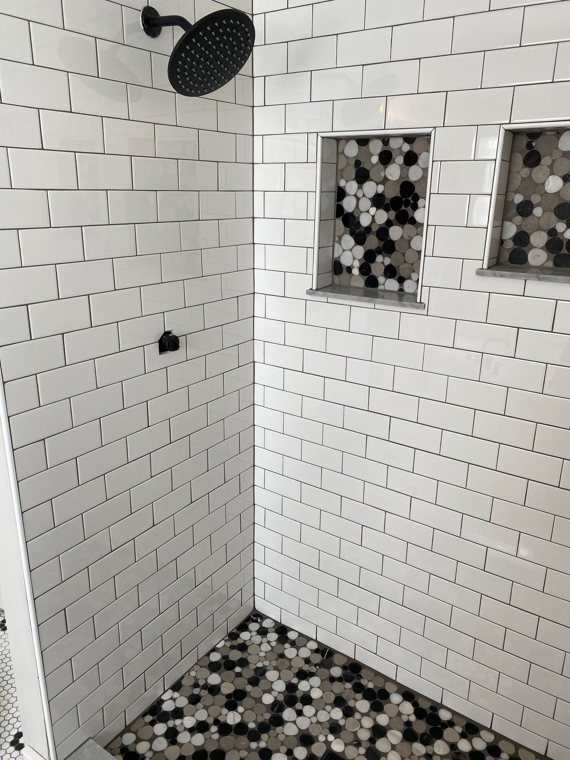 Small white-tiled bathroom shower with black fixtures and pebble-style floor tiles.