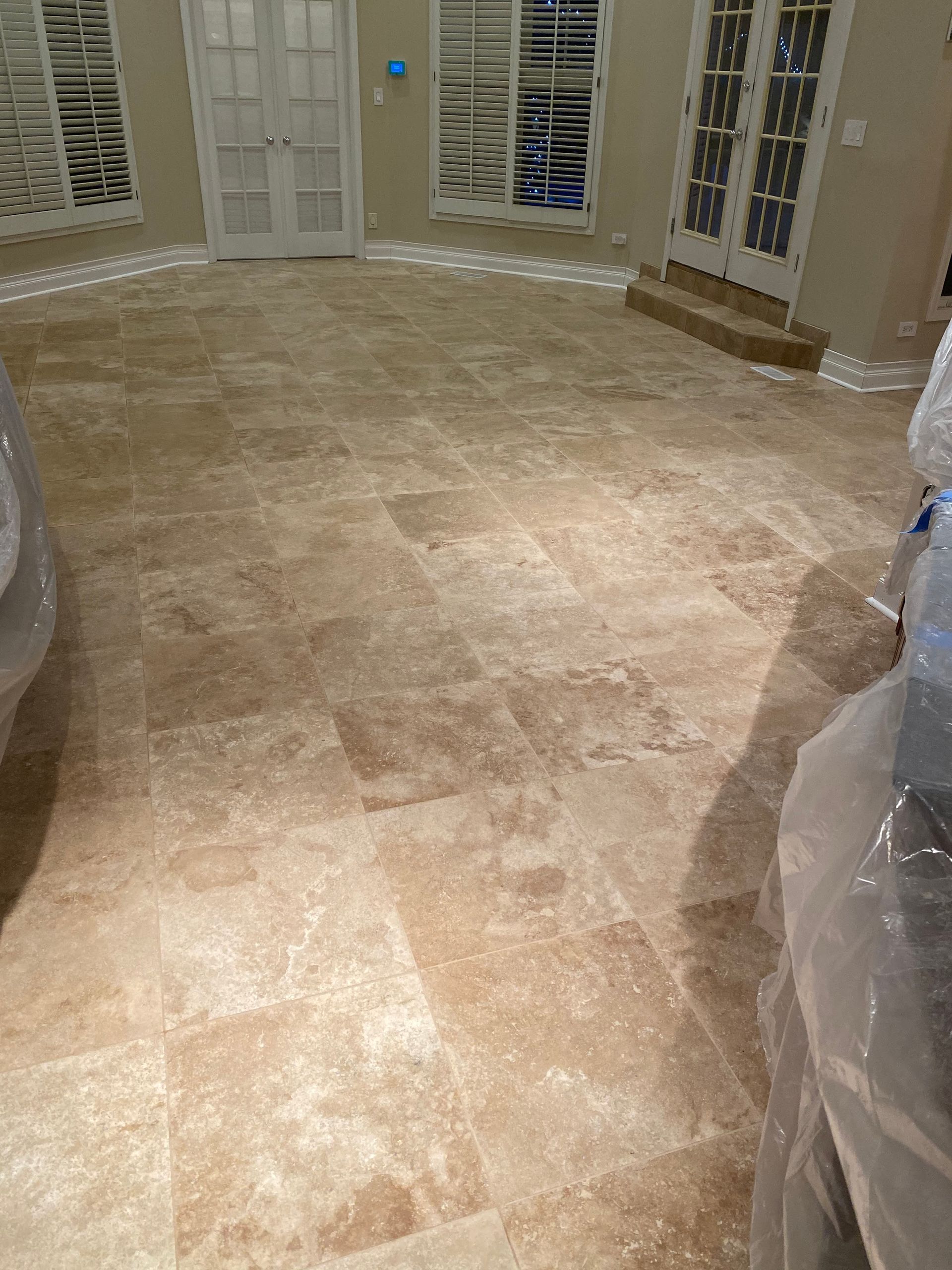 Empty tiled living room with white French doors and covered furniture at the edges