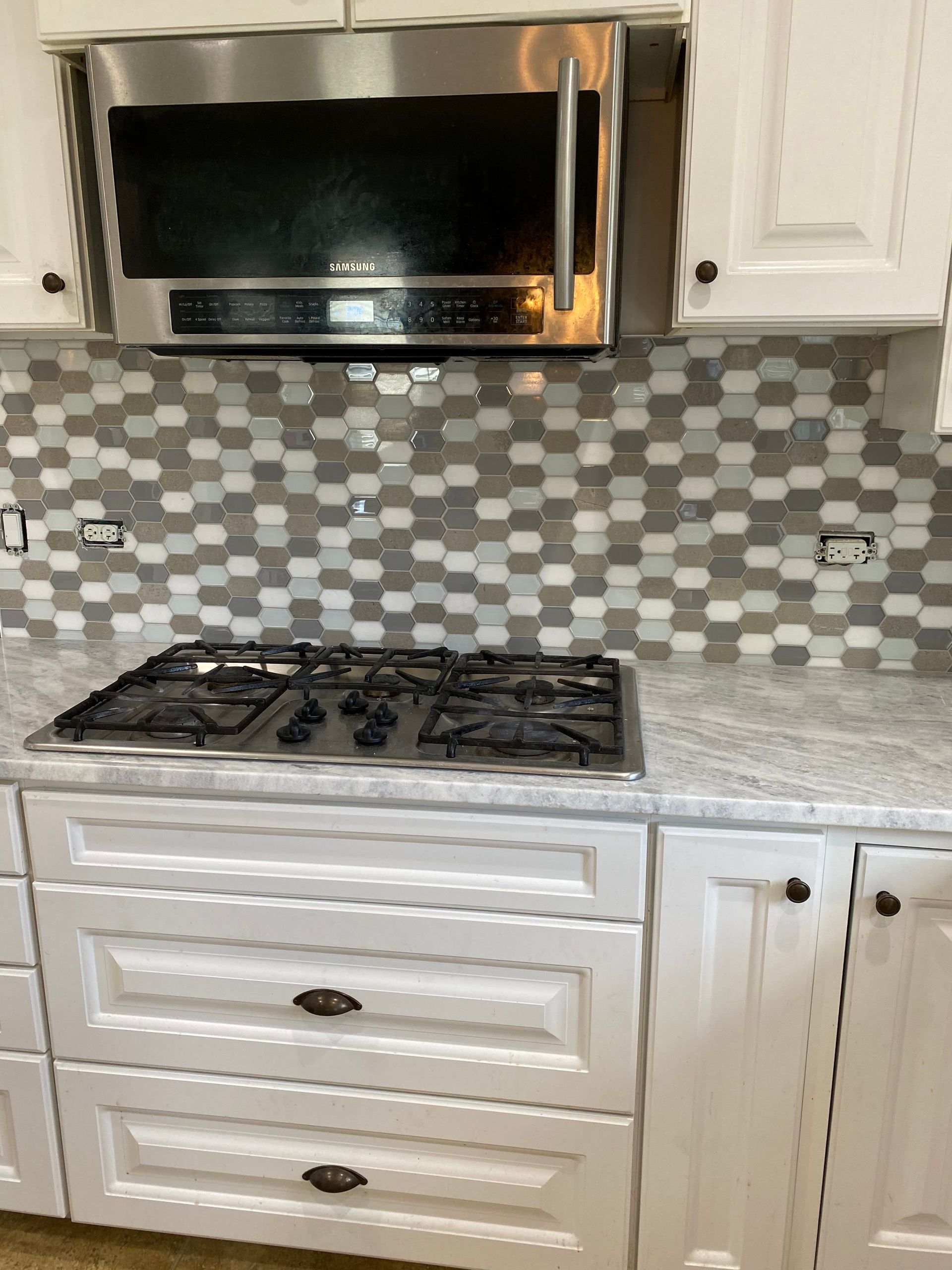 Kitchen with white cabinets, stainless steel microwave, gas stovetop, and gray mosaic tile backsplash.