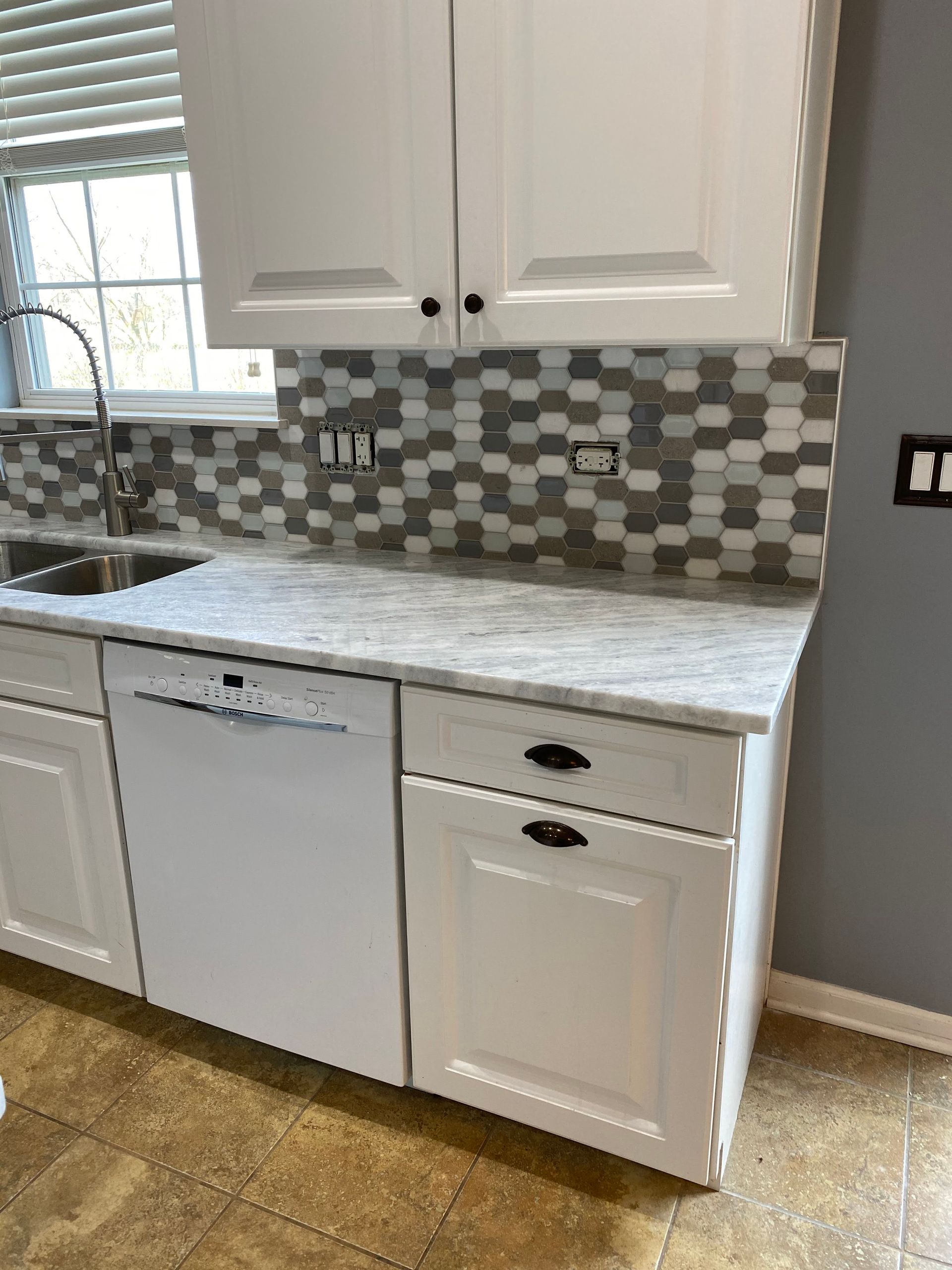 White kitchen cabinets and dishwasher with gray mosaic backsplash and countertop by a window