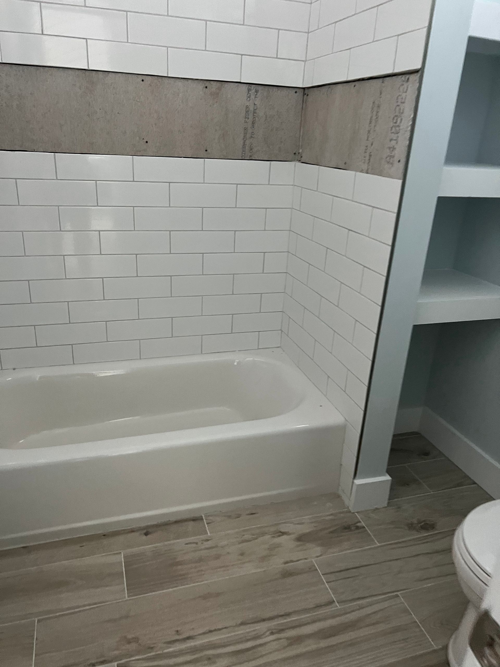 White bathtub in a tiled bathroom, with a gray accent band and an open shelving unit nearby.