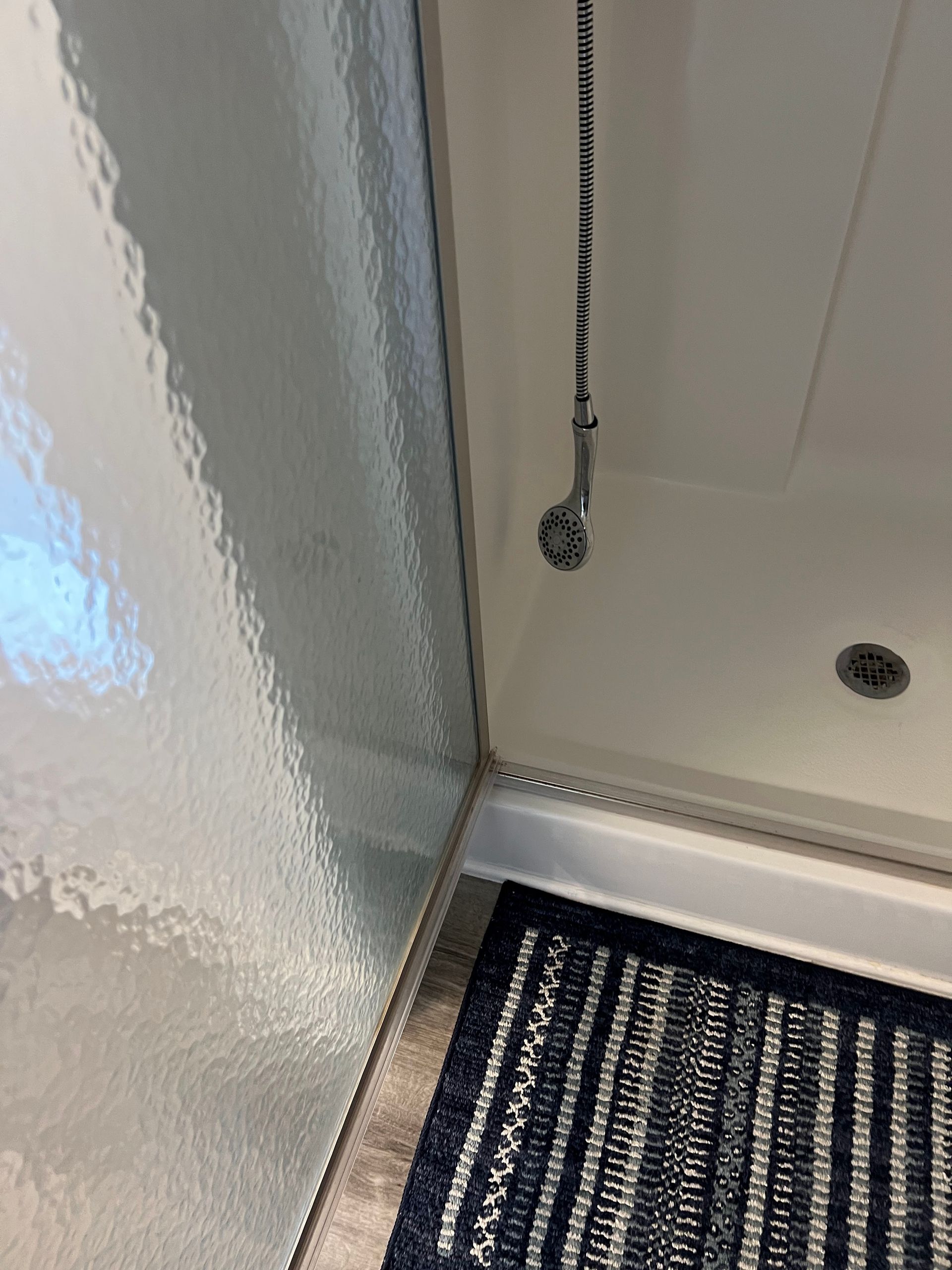Shower stall corner with a pull chain, chrome trim, and a striped bath mat below.