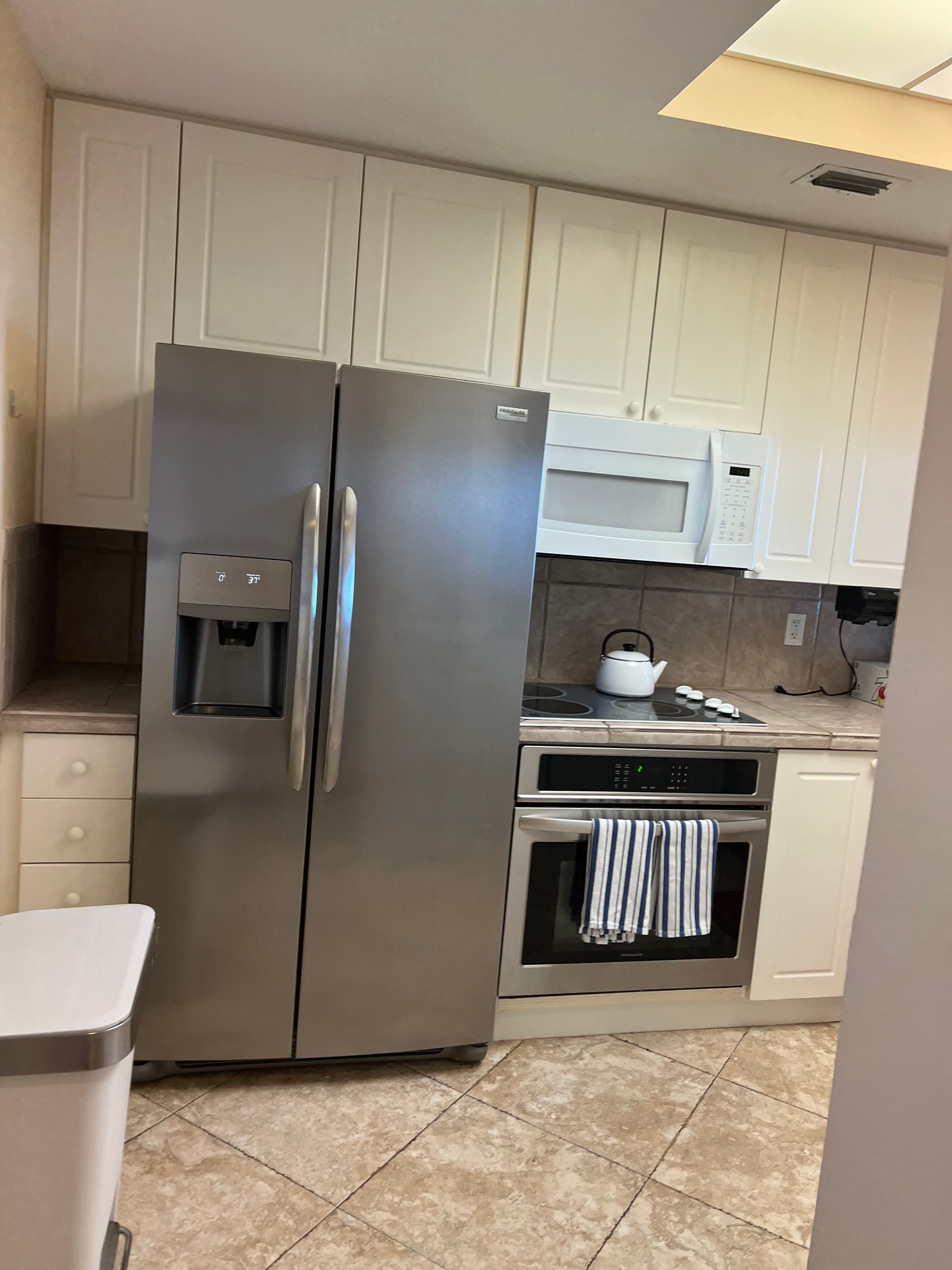 Kitchen with stainless steel refrigerator, white cabinets, stove, and beige tiled floor.