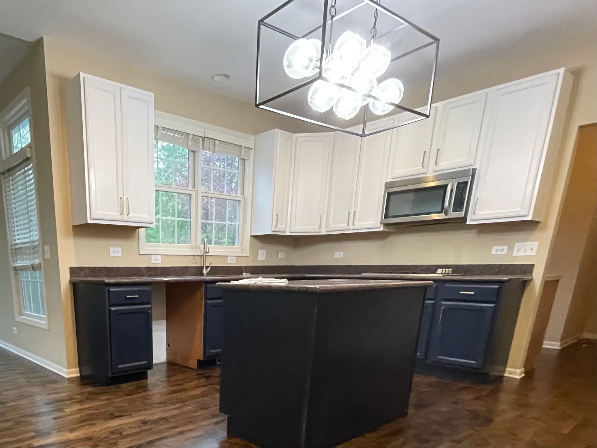 Modern kitchen with white cabinets, dark island, stainless microwave, and large window.