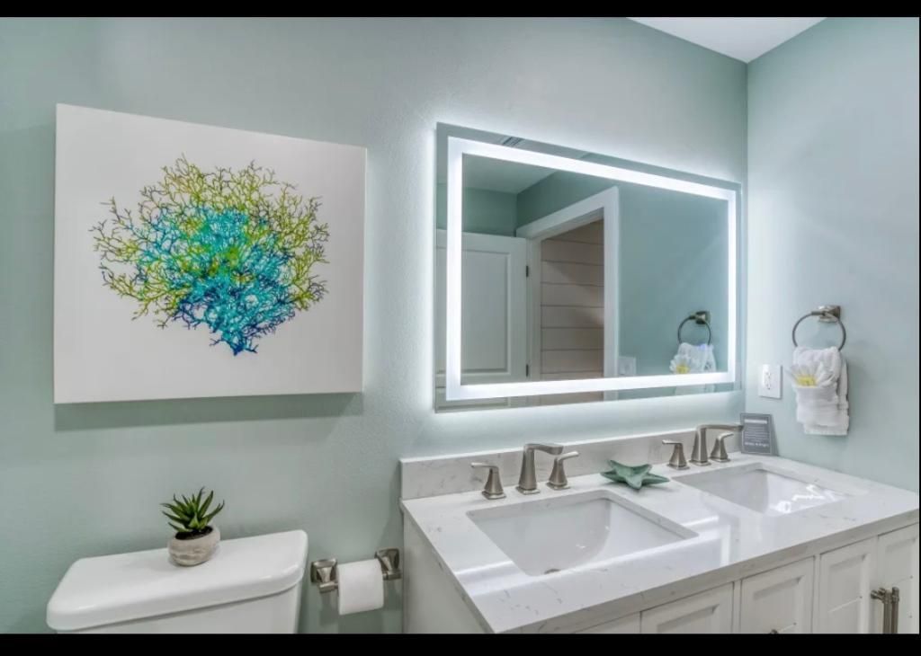 Modern bathroom with double sink, illuminated mirror, white cabinetry, and abstract wall art