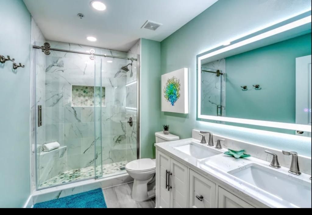 Bright teal bathroom with glass shower, double vanity, and large lit mirror