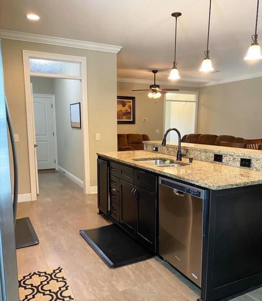 Modern kitchen with granite island, pendant lights, stainless dishwasher, and hallway visible.