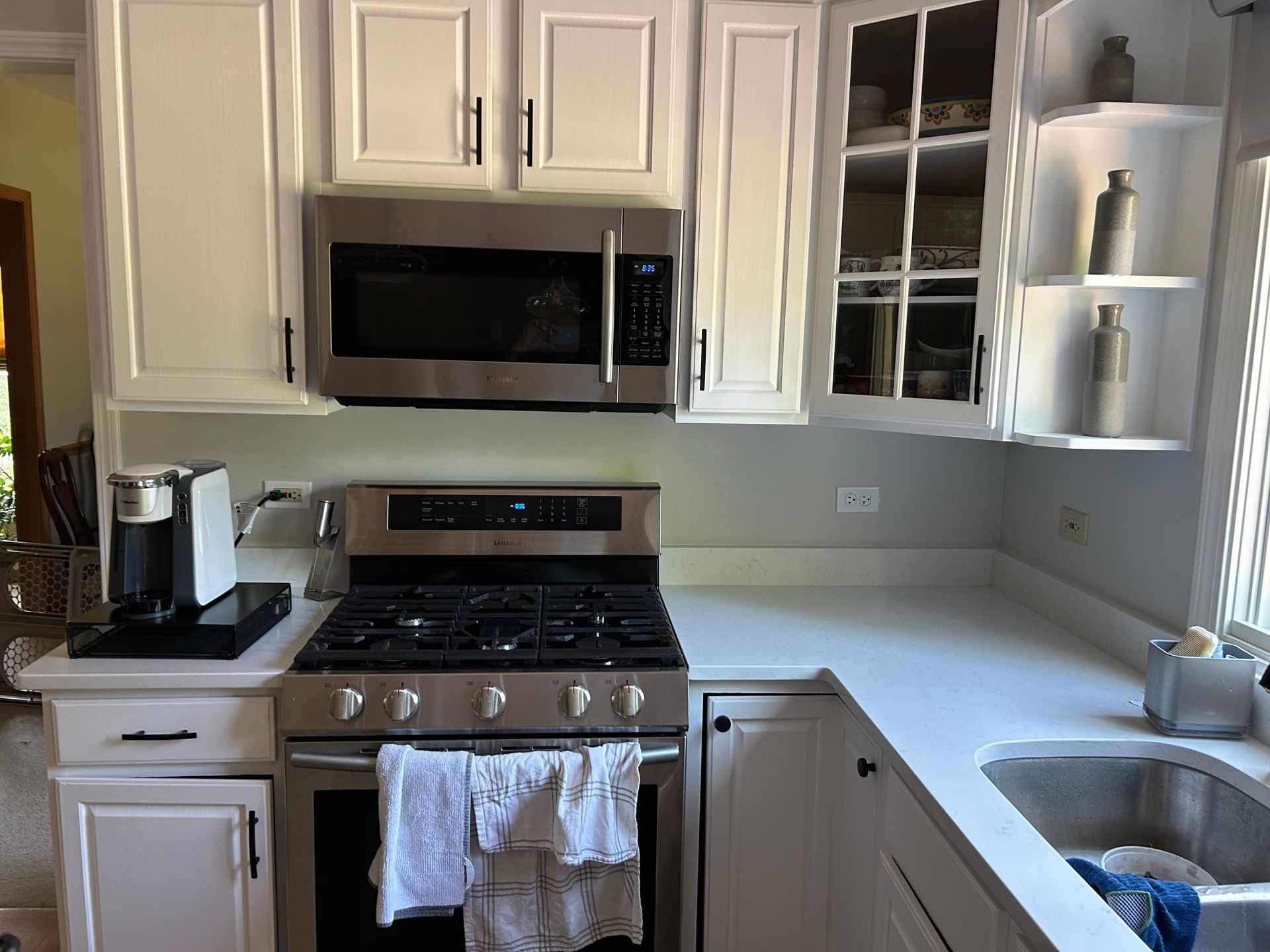 White kitchen with stainless stove and microwave, cabinets, countertop, and sink by a window.