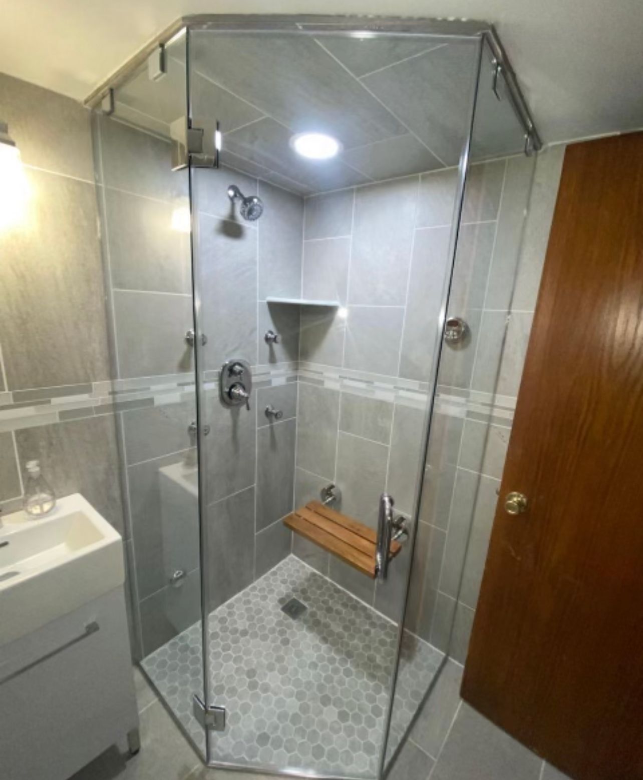 Glass-enclosed corner shower with gray tile, wall shelf, and wooden shower bench in a bathroom