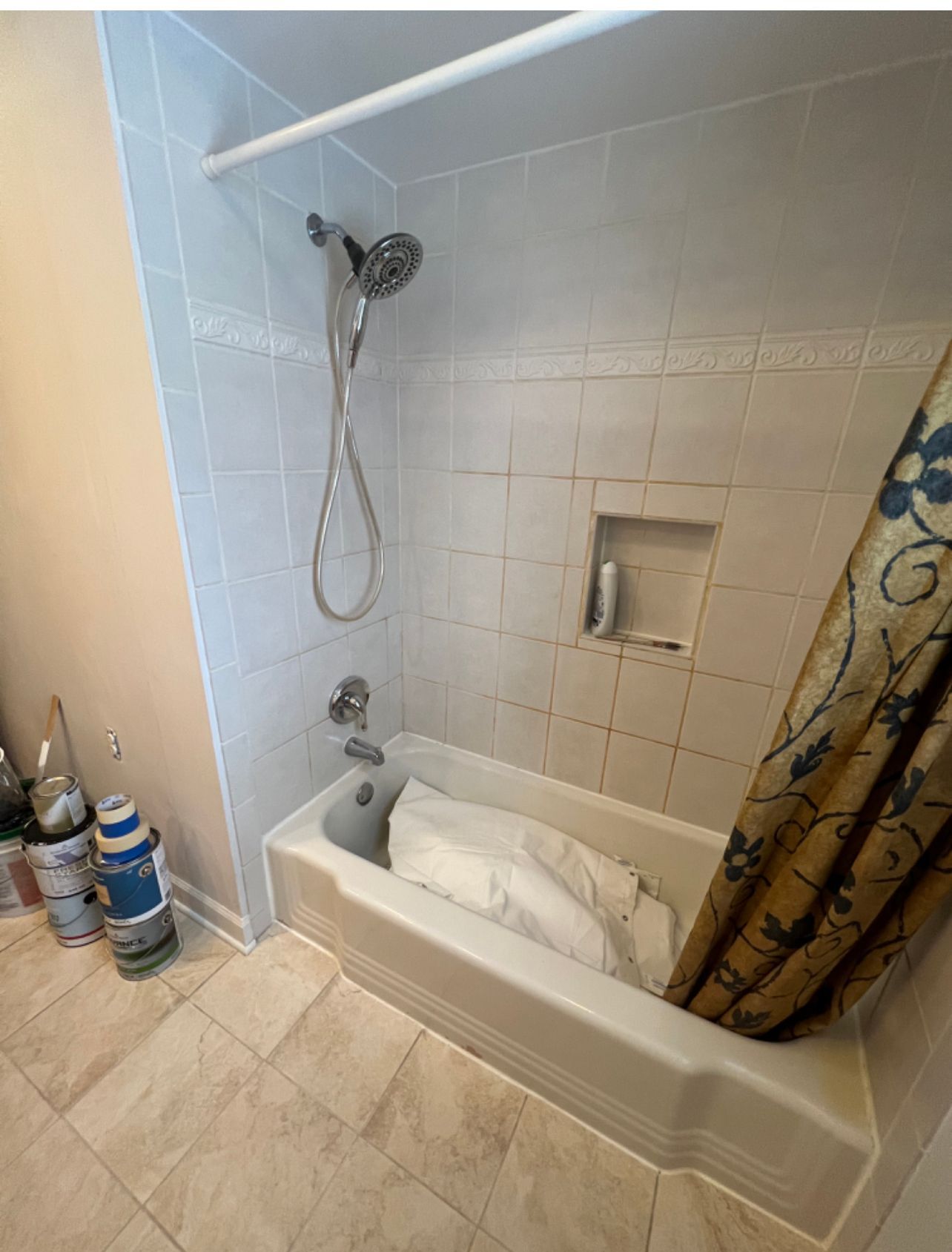 Small tiled bathroom with a shower-tub combo, beige curtains, and cleaning supplies on the floor.