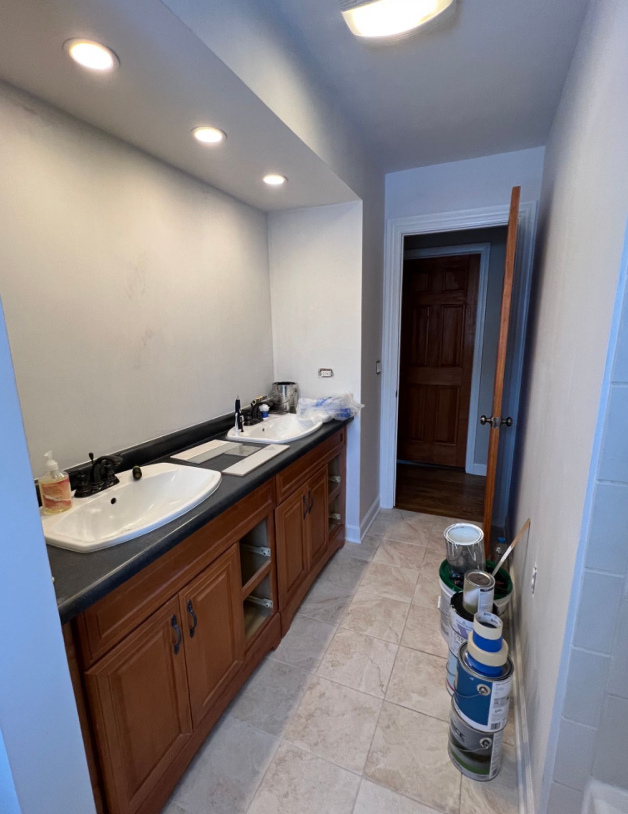 Narrow bathroom with double sink vanity, recessed lights, and an open doorway at the end.