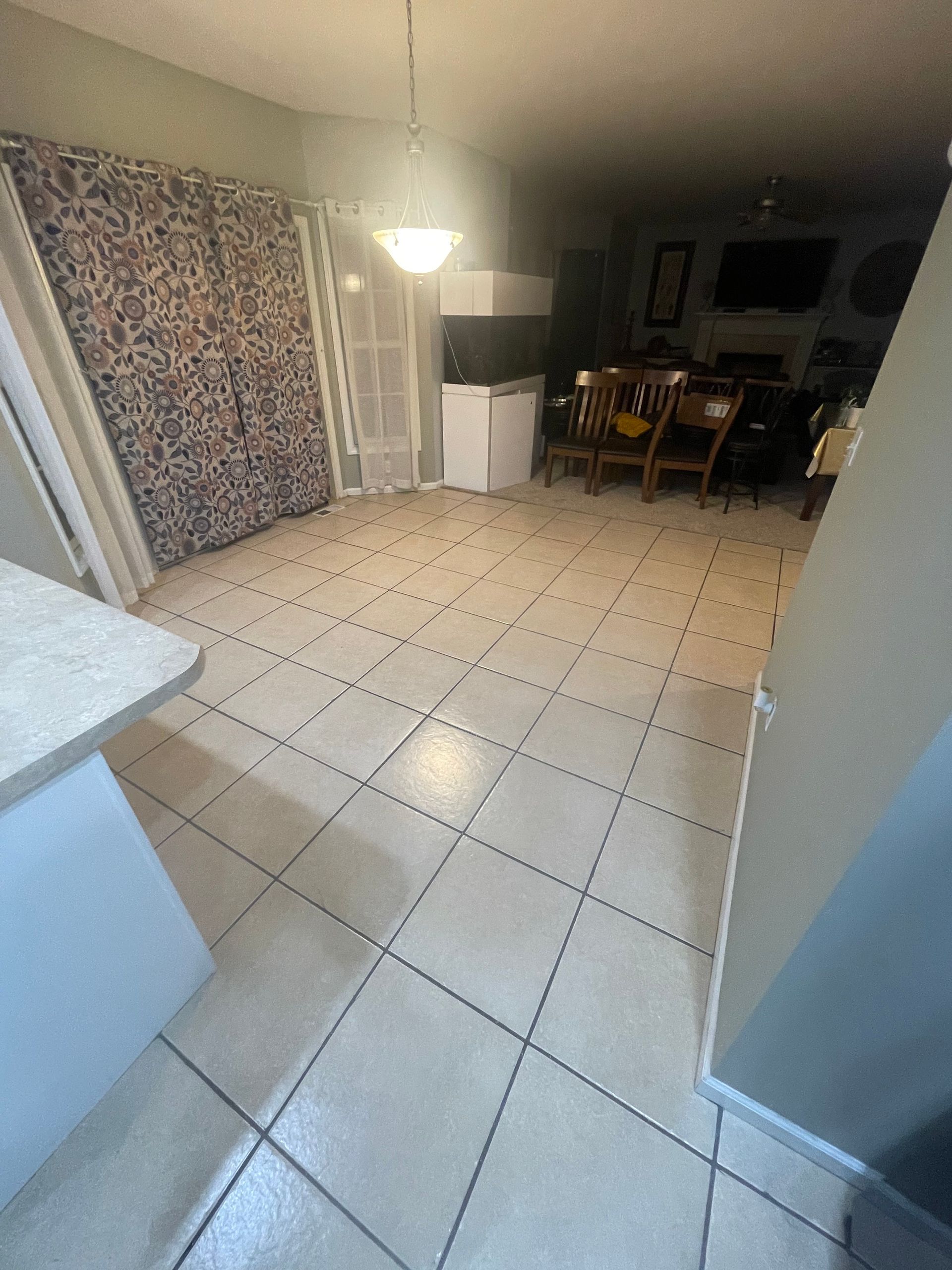 Empty tiled living room with a folded mattress leaning against the wall and a small dining area in the back.