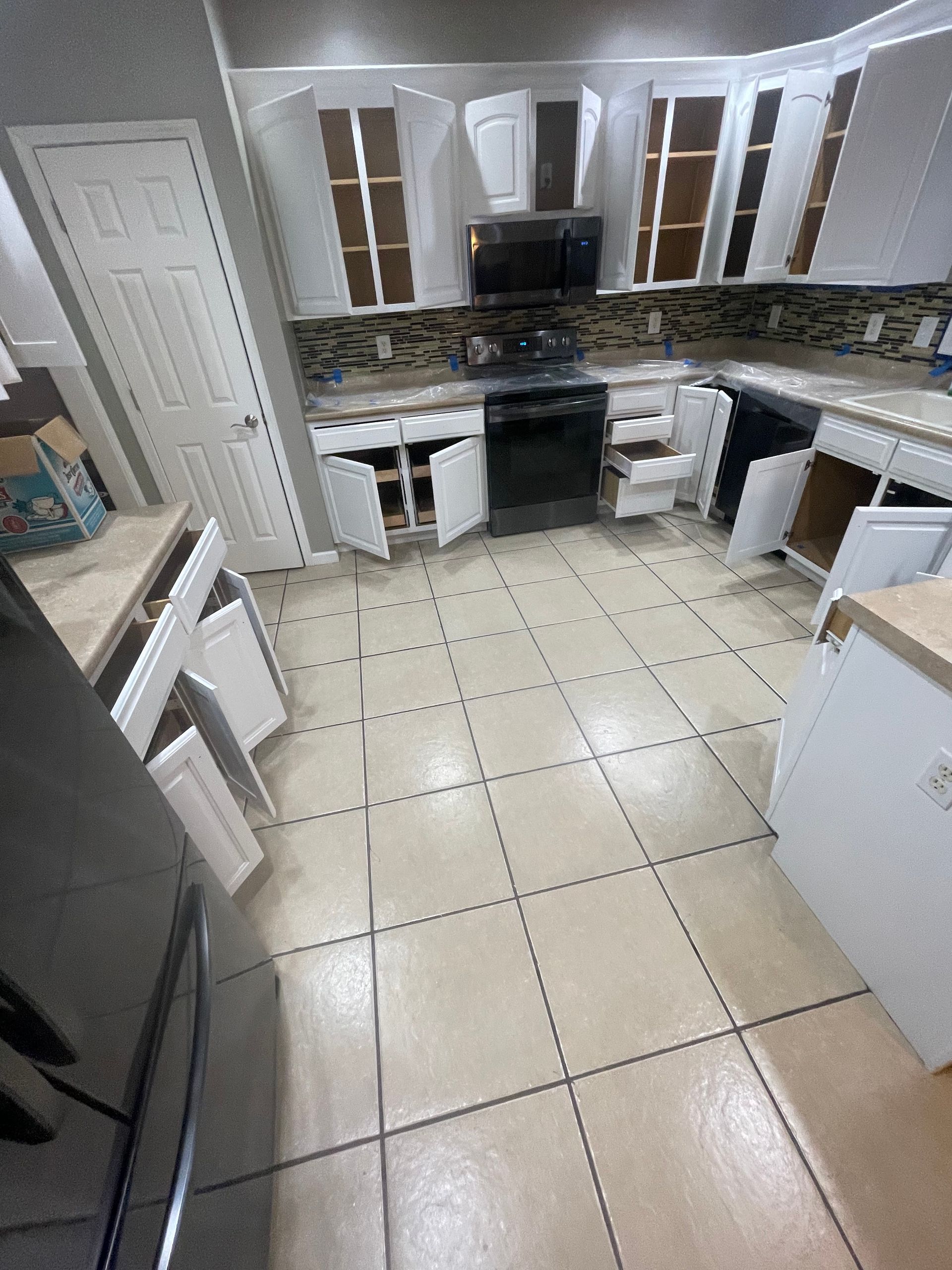 Empty kitchen with beige tile floor, white cabinets, black stove, and windows above the sink.