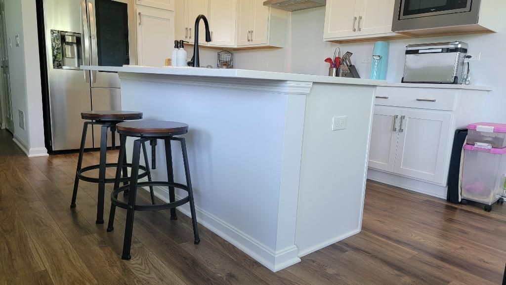 Open kitchen with white cabinets, island, and two dark stools on wood flooring.