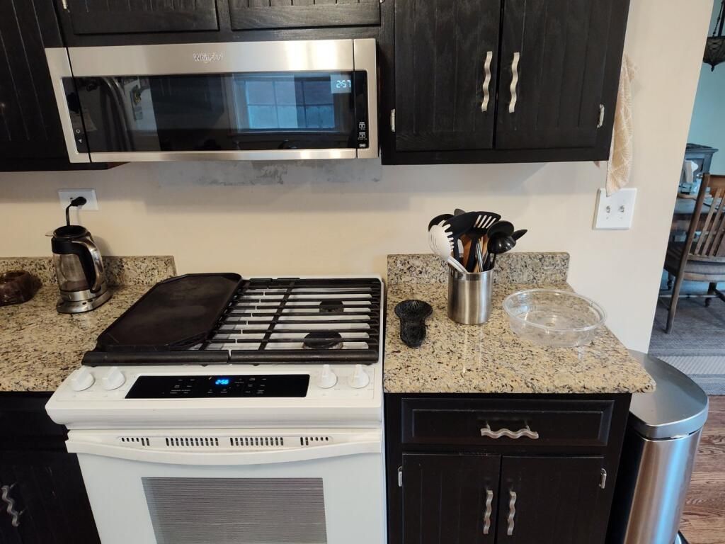 Kitchen with white stove, microwave, granite countertop, dark cabinets, and utensils on the counter