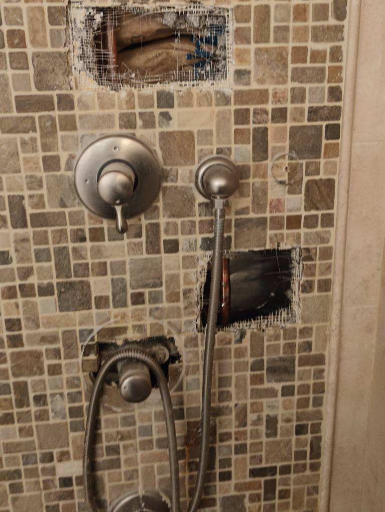 Shower wall with two metal controls, handheld sprayer, and tiled beige mosaic backdrop