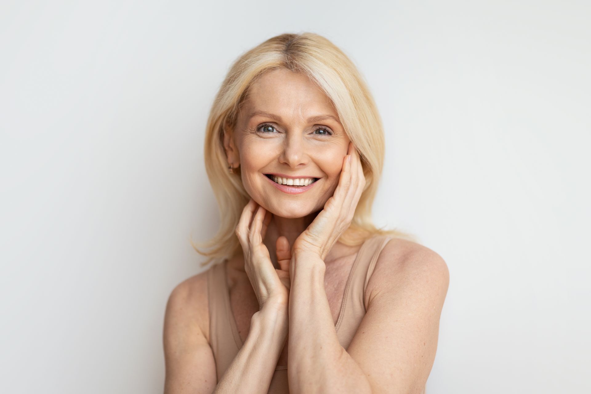 Smiling blonde woman touching her face against a light gray background