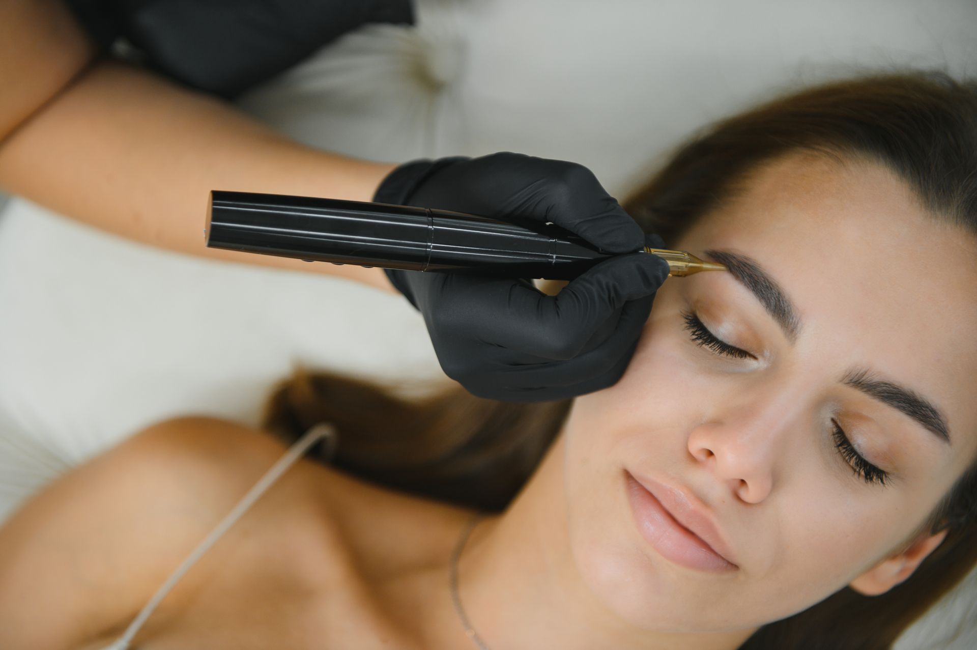 Gloved technician applying treatment to a woman’s eyebrow with a fine tool, close-up.