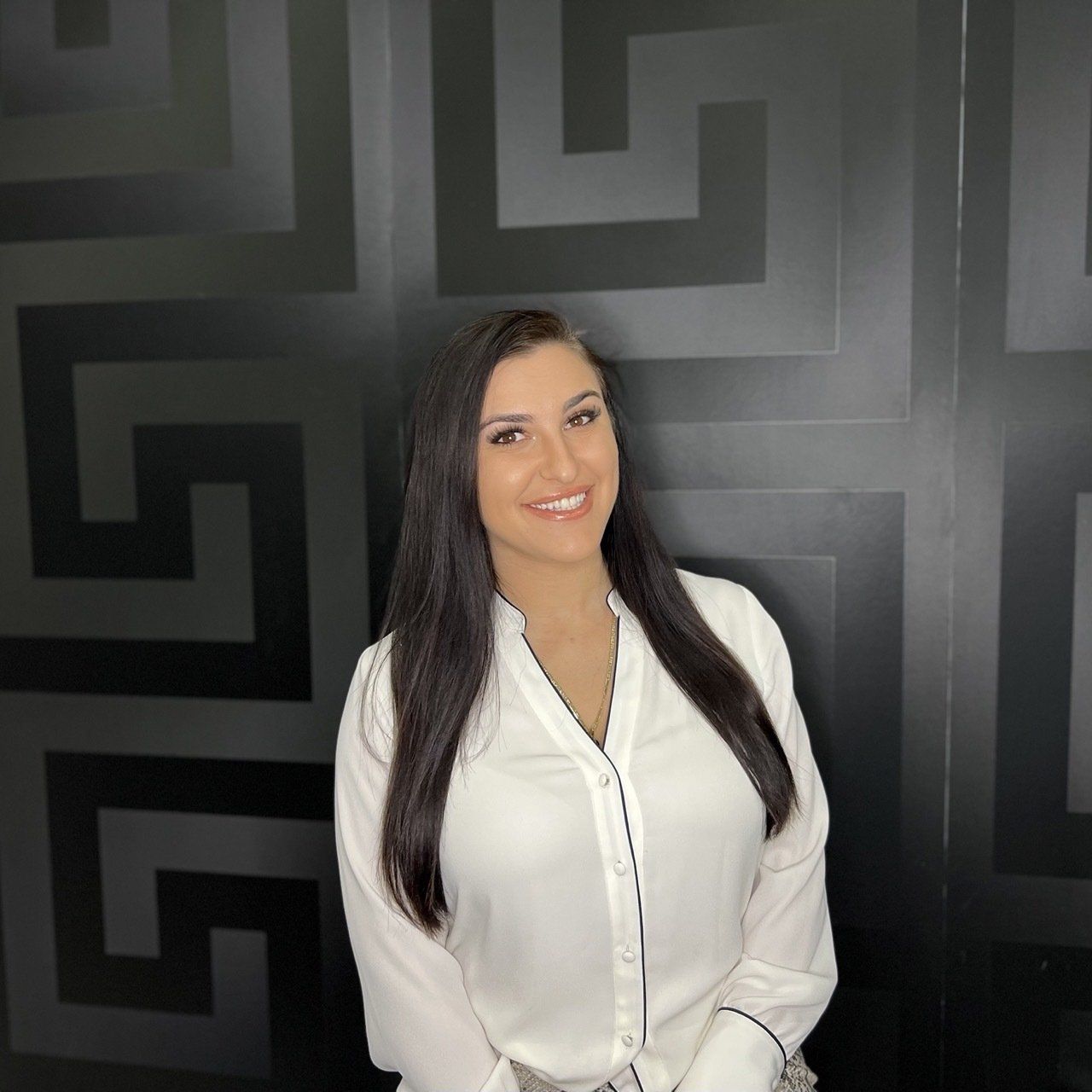 Woman smiling, wearing white button-down shirt, standing in front of a black geometric pattern wall.