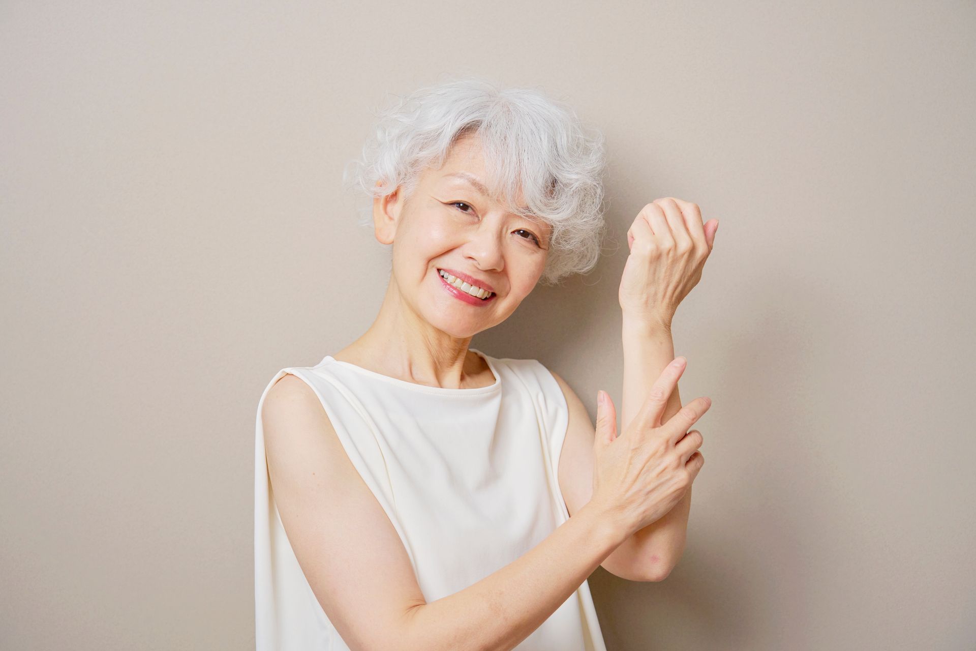 Woman smiles, touches arm, white top, short gray hair, against a neutral background.