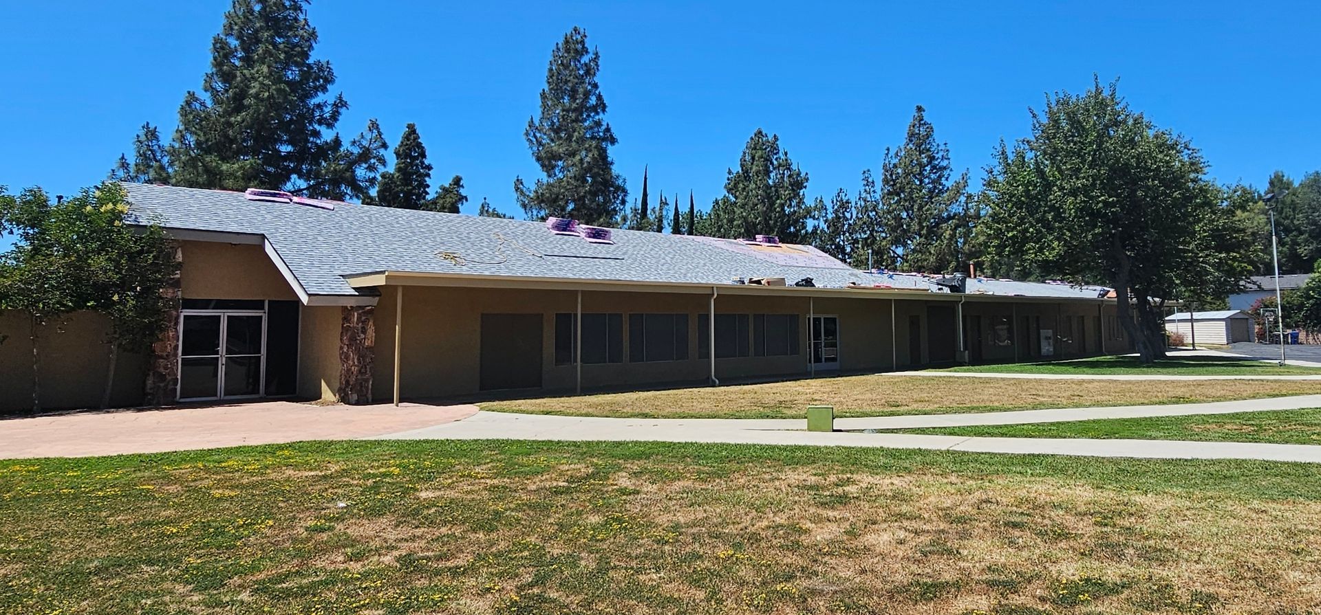 A one-story beige building with a gray roof under a clear blue sky, with trees and grass in the foreground.