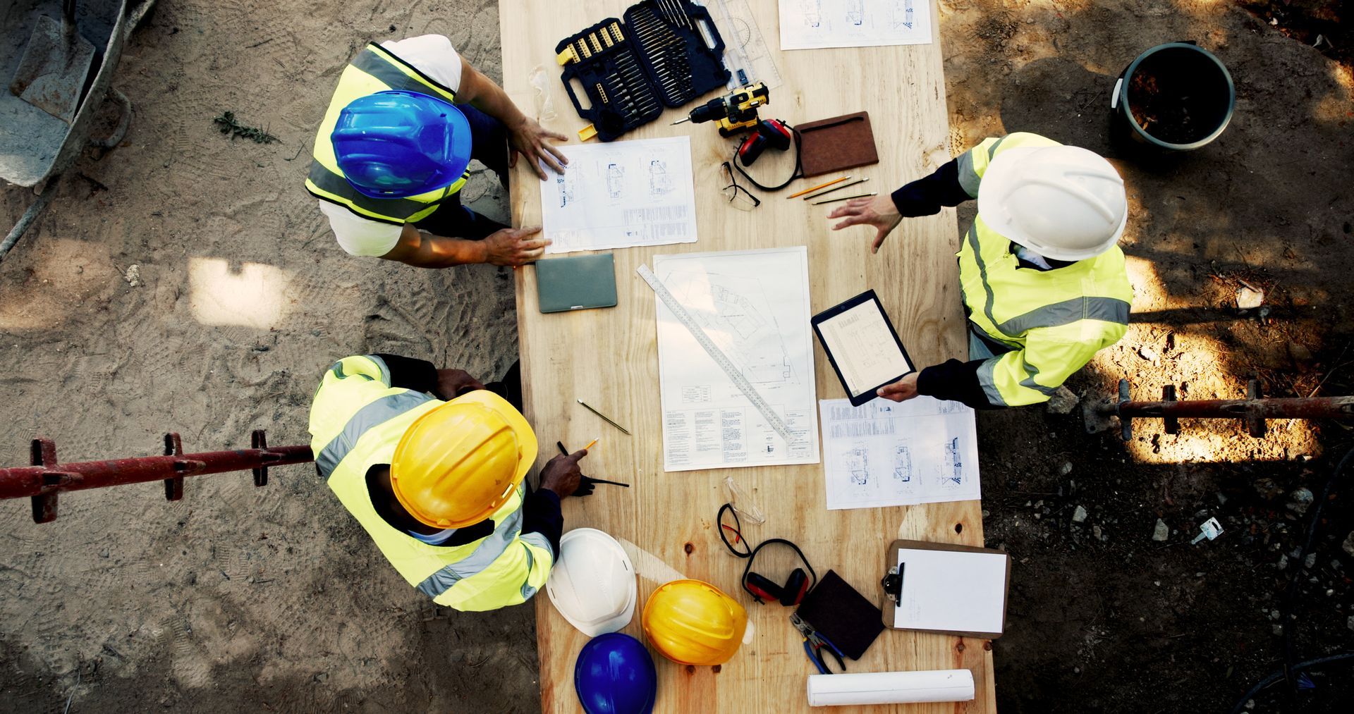 Three construction workers in hard hats and safety vests review blueprints and a tablet on a table at a job site.