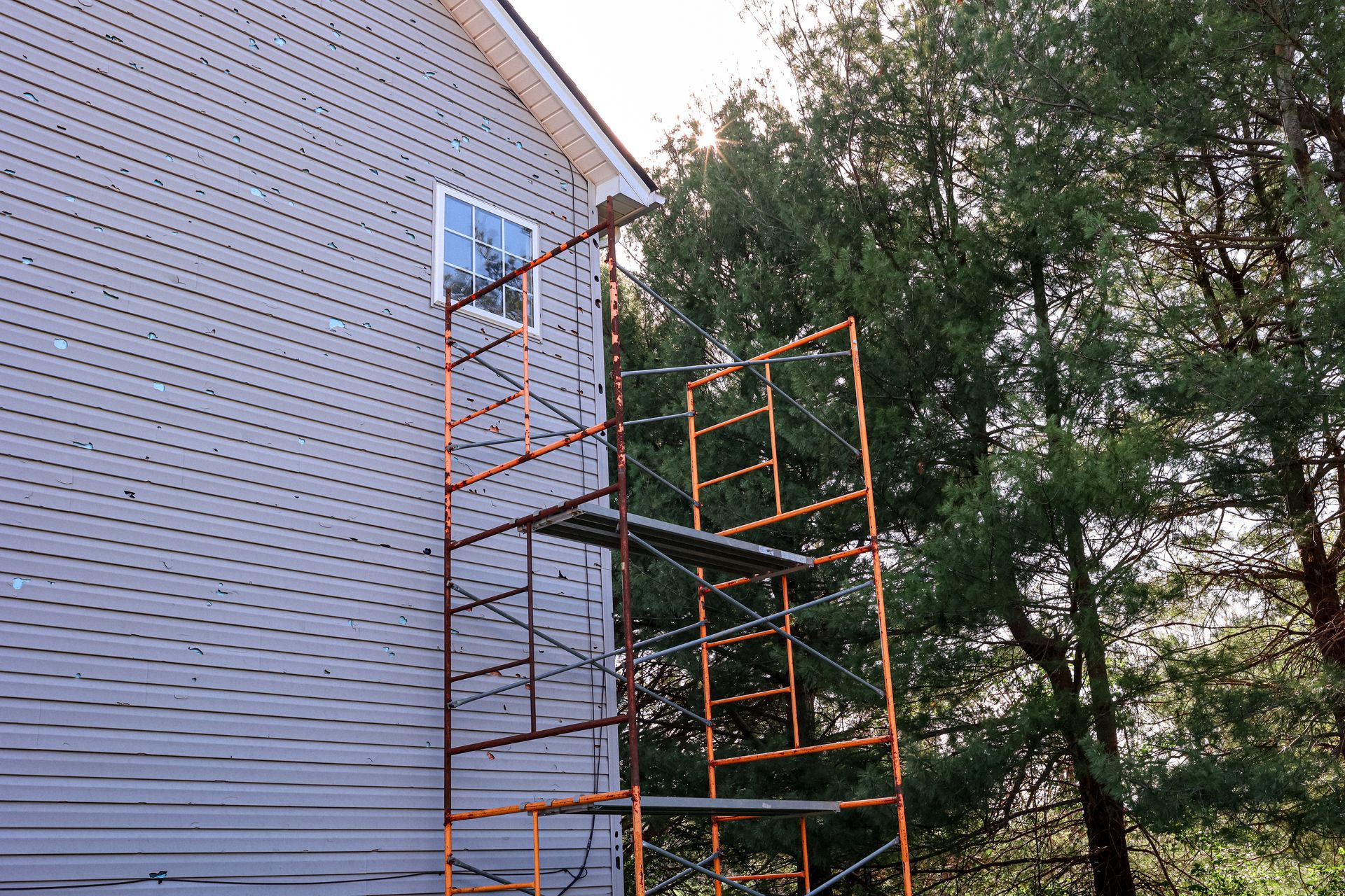 Un échafaudage métallique orange est installé contre le côté d'une maison aux murs gris, à côté d'une fenêtre et d'arbres.