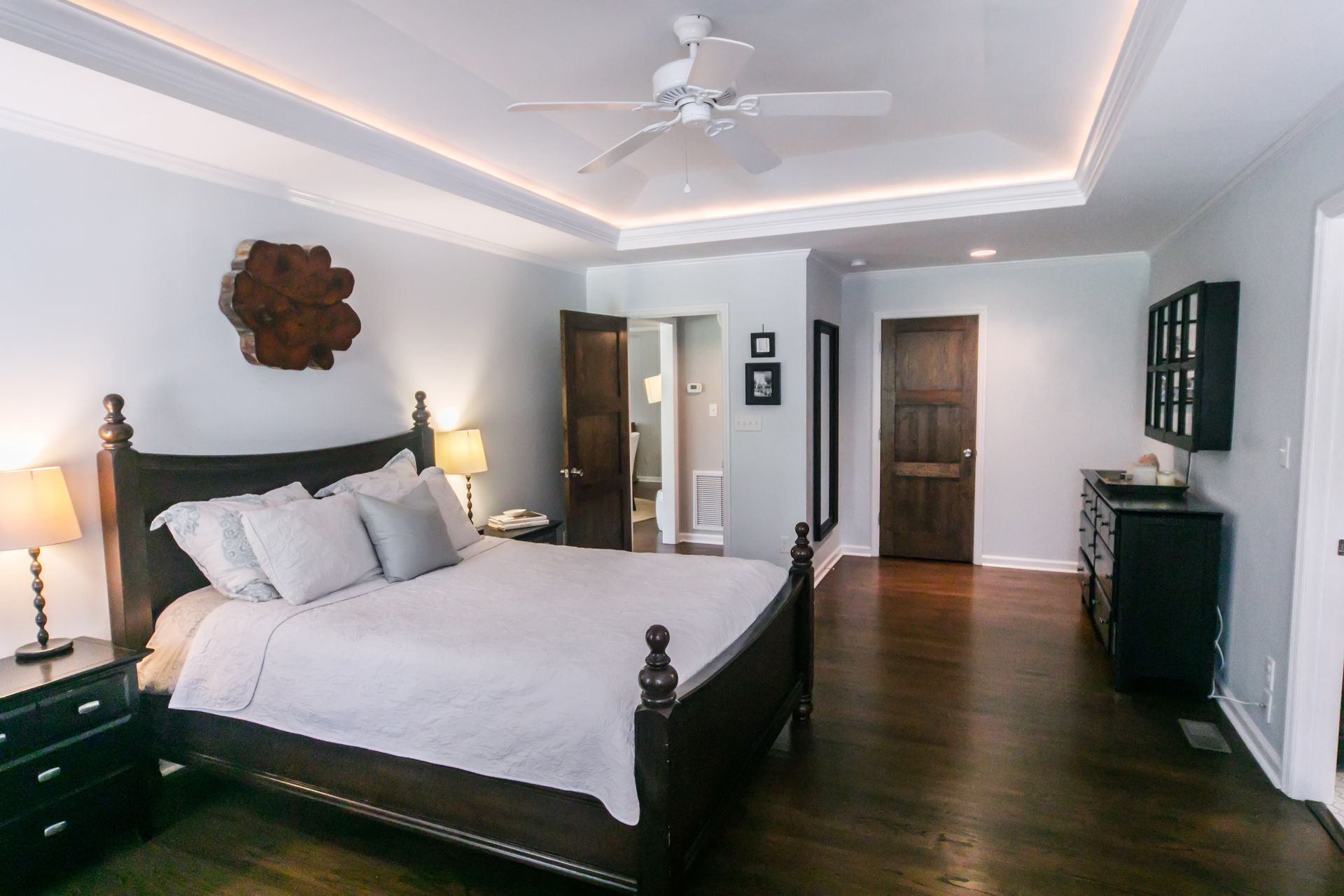 Une chambre spacieuse avec un lit en bois foncé, des draps blancs, du parquet, un ventilateur de plafond et des portes en bois foncé.