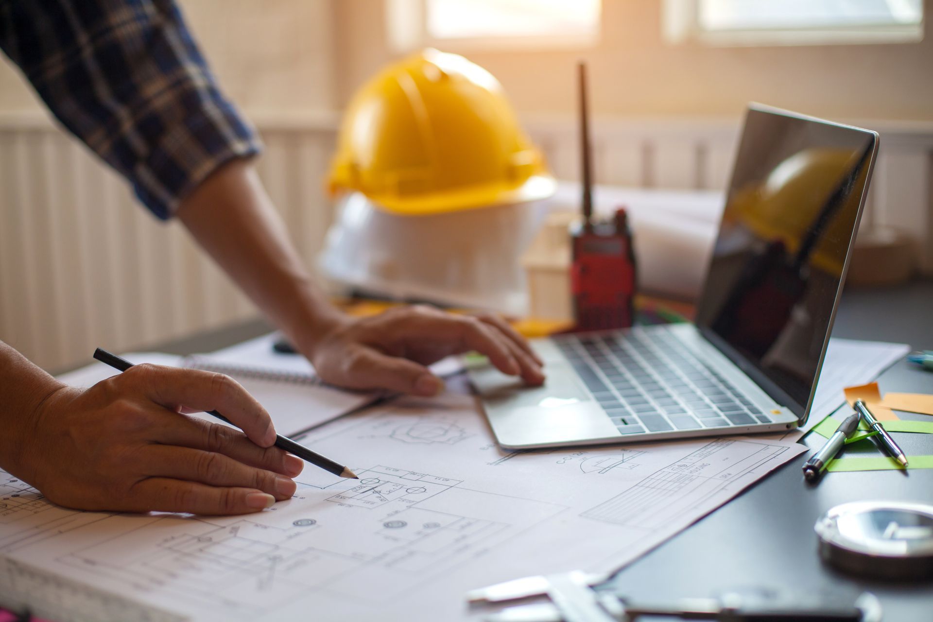 Les mains d'un ingénieur tenant un crayon au-dessus de plans, à côté d'un ordinateur portable et d'un casque de chantier jaune posés sur un bureau.