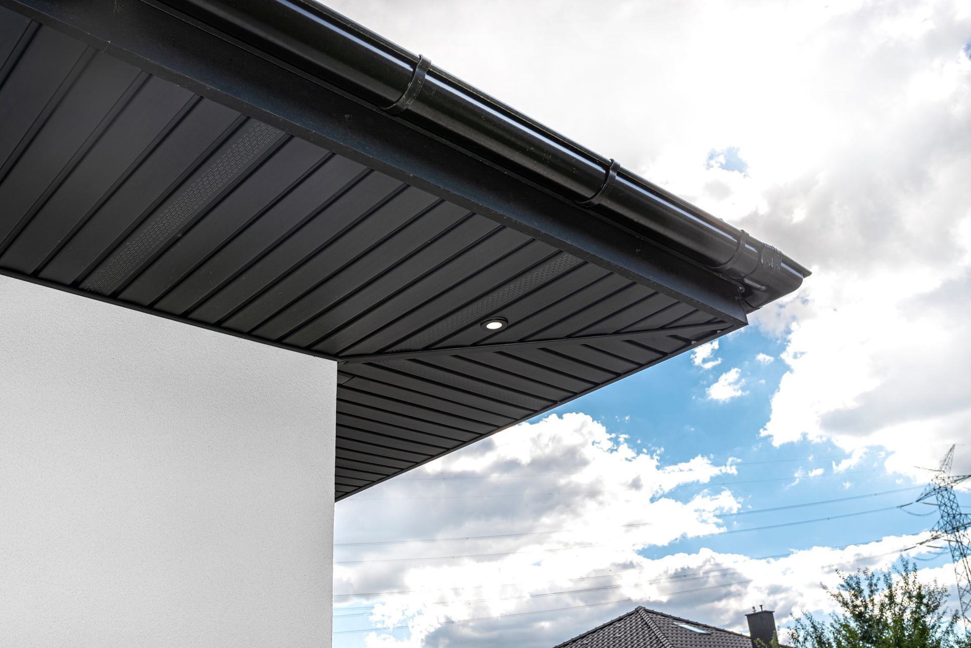 Low-angle view of a modern house's black exterior soffit, gutter, and roofline against a cloudy blue sky.