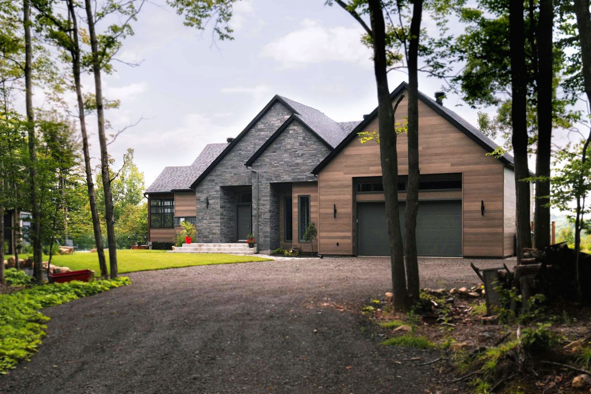 Une maison moderne aux façades en pierre grise et en bois, située dans un paysage boisé avec une allée de gravier menant à un garage.