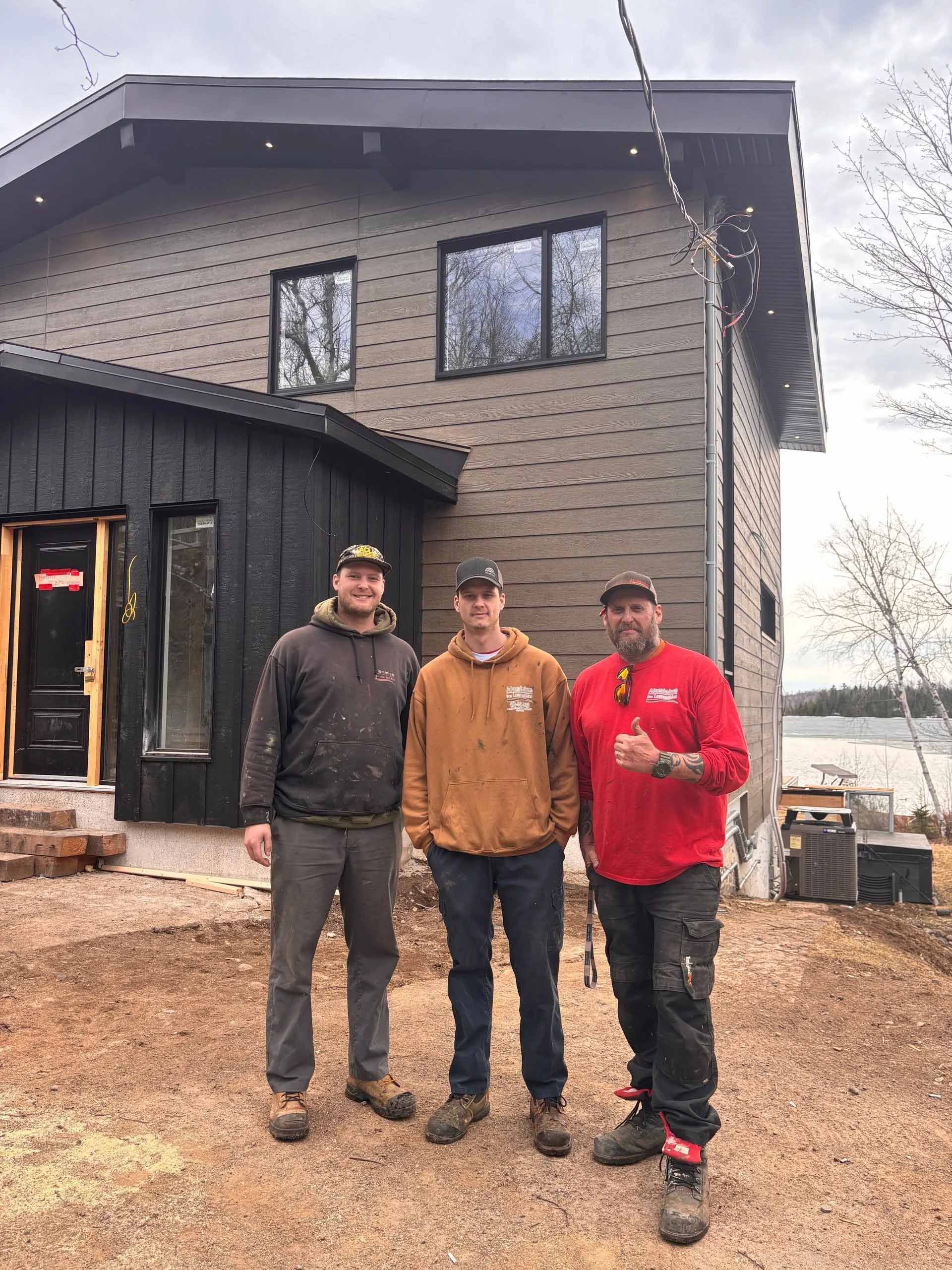 Three people standing in front of a modern house under construction with brown patterned siding and a black entryway.