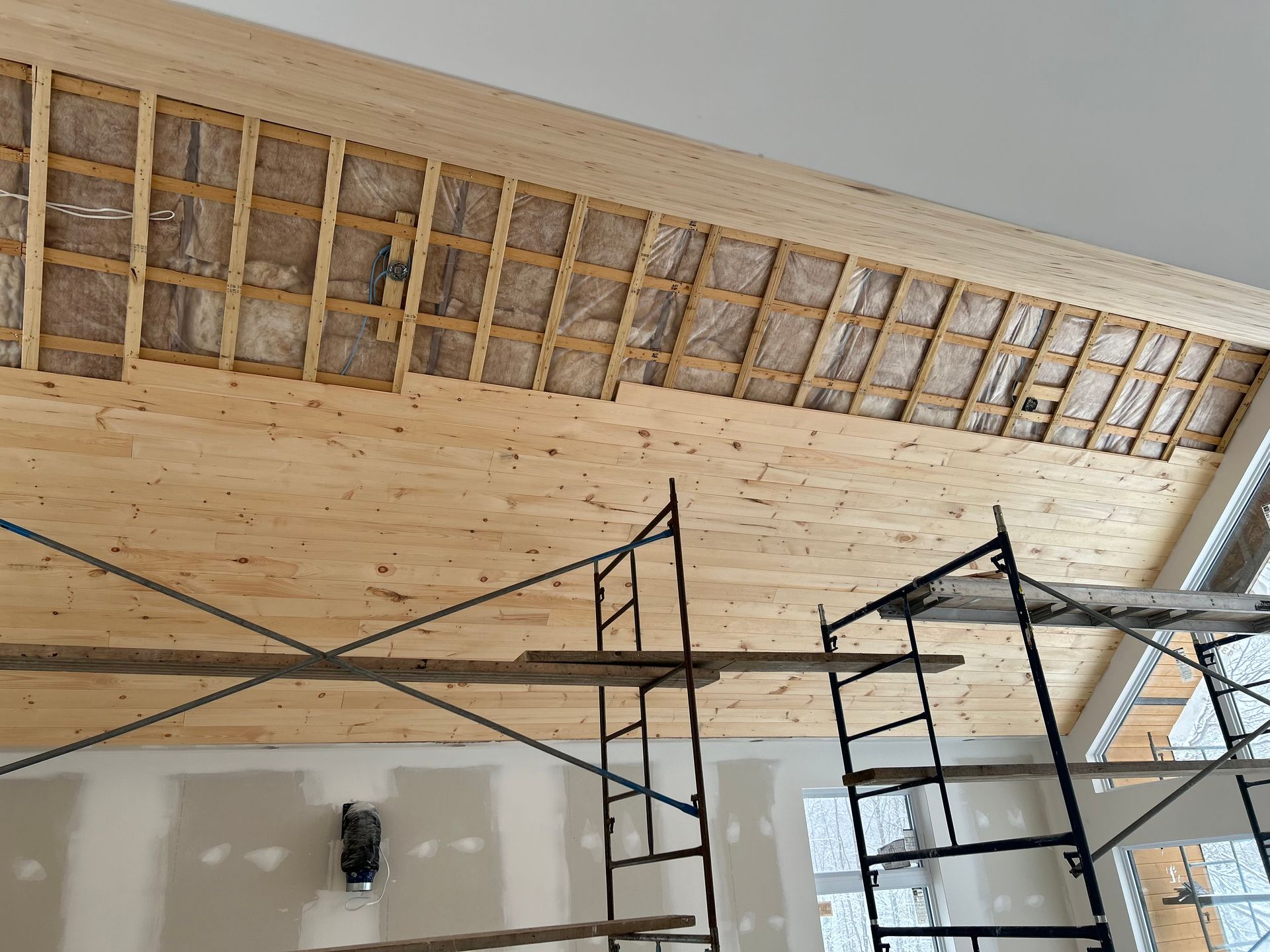 Vue intérieure d'un chantier montrant les chevrons de plafond en bois apparents avec isolation, des planches partiellement finies et un échafaudage.