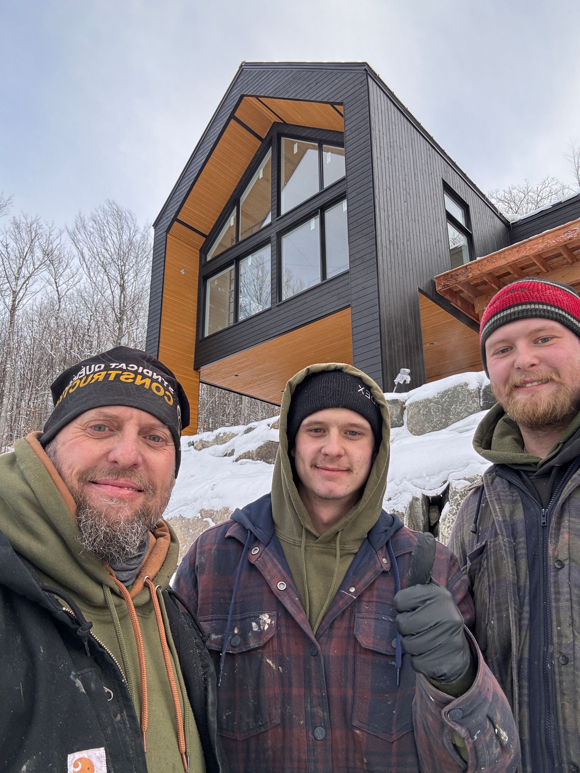 Trois personnes en vêtements d'hiver posent devant un chalet moderne aux lignes angulaires, avec un revêtement en métal noir et des éléments en bois.