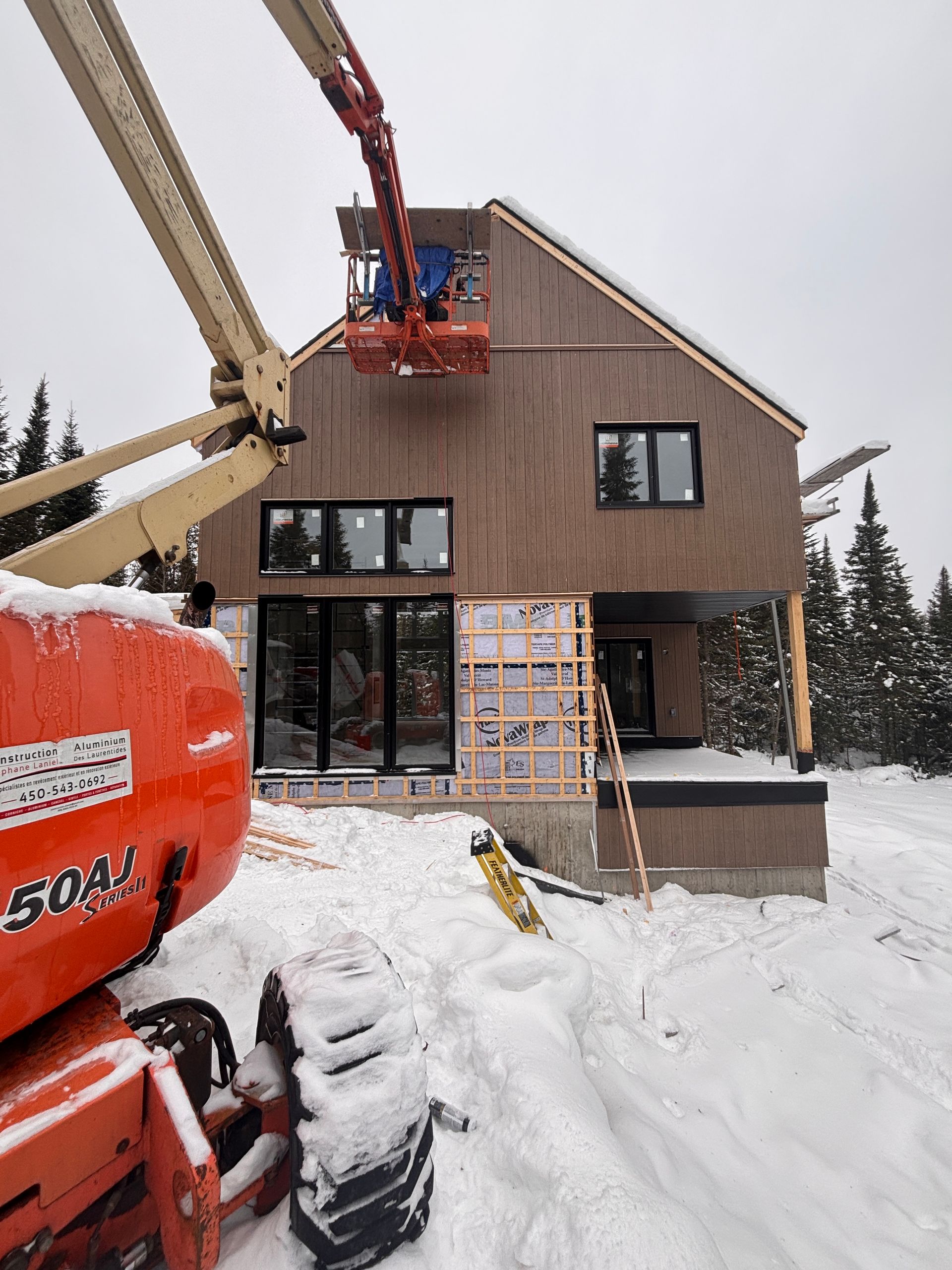Un monte-charge de chantier atteint le niveau supérieur d'une maison brune en construction dans un décor forestier enneigé.