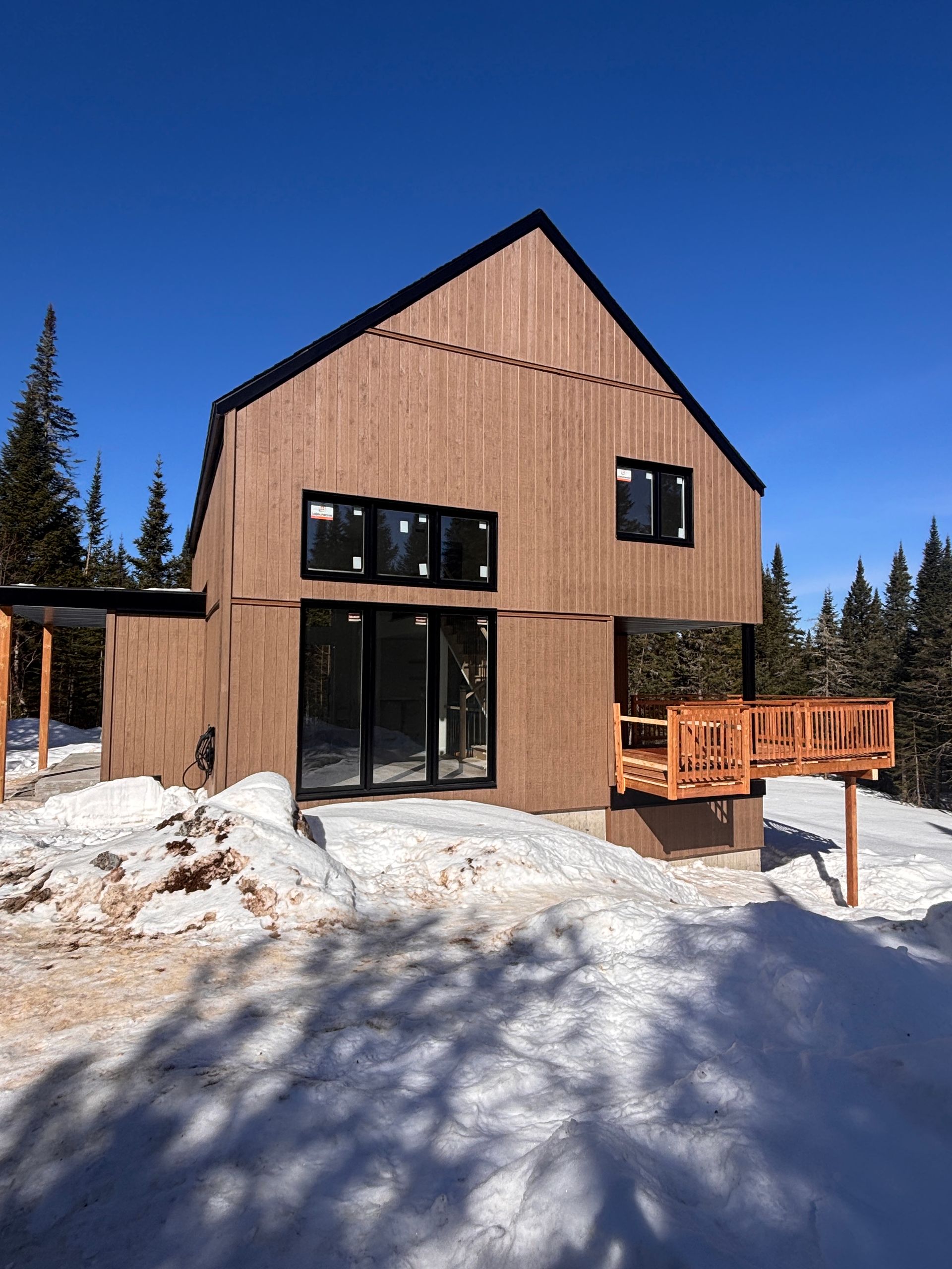 Une cabane moderne en bois, à la structure verticale, se dresse dans un paysage montagneux enneigé, sous un ciel d'un bleu limpide.