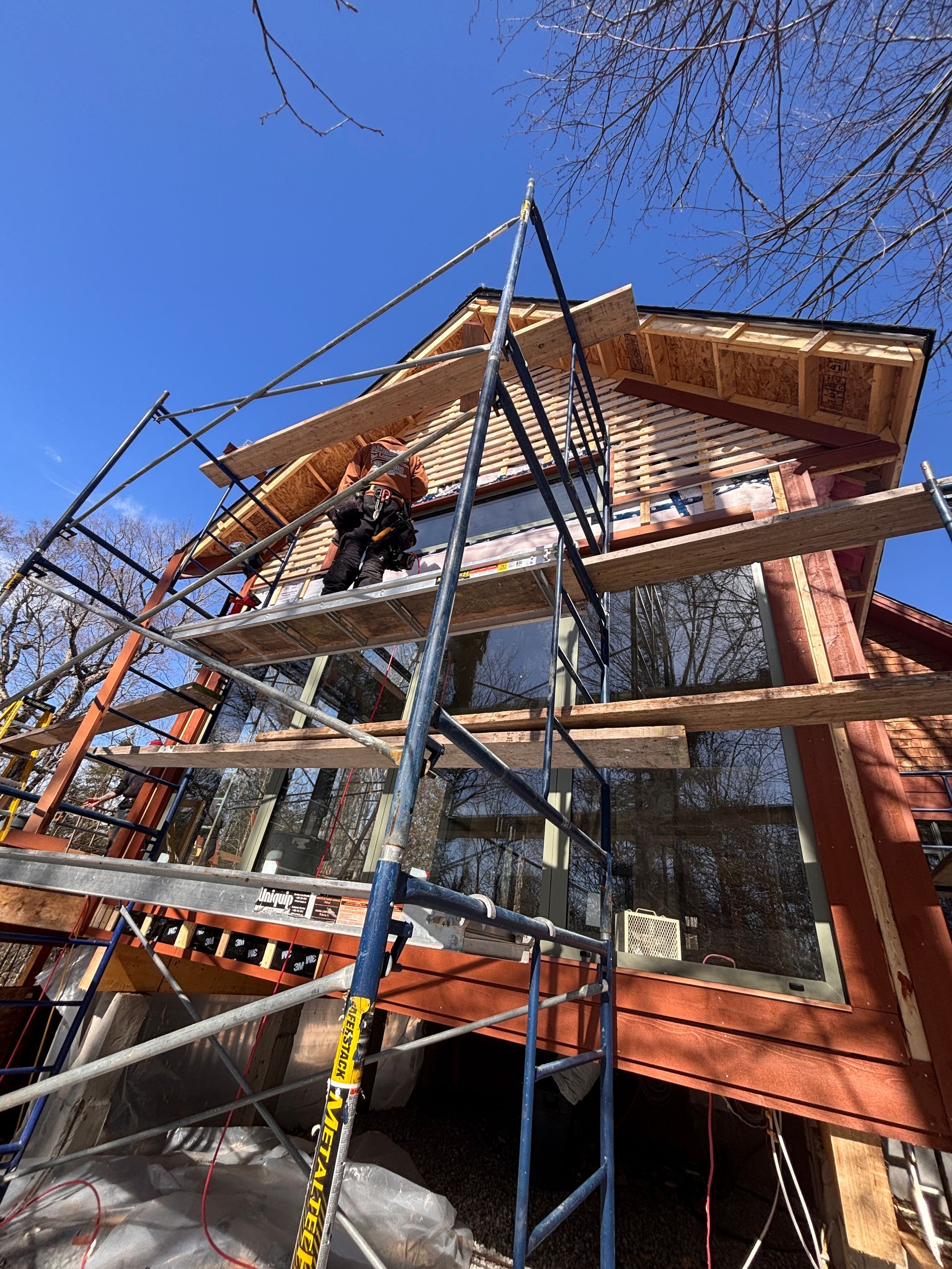 Sous un ciel d'un bleu éclatant, un ouvrier du bâtiment, juché sur un échafaudage, installe un bardage en bois sur la façade d'une maison.