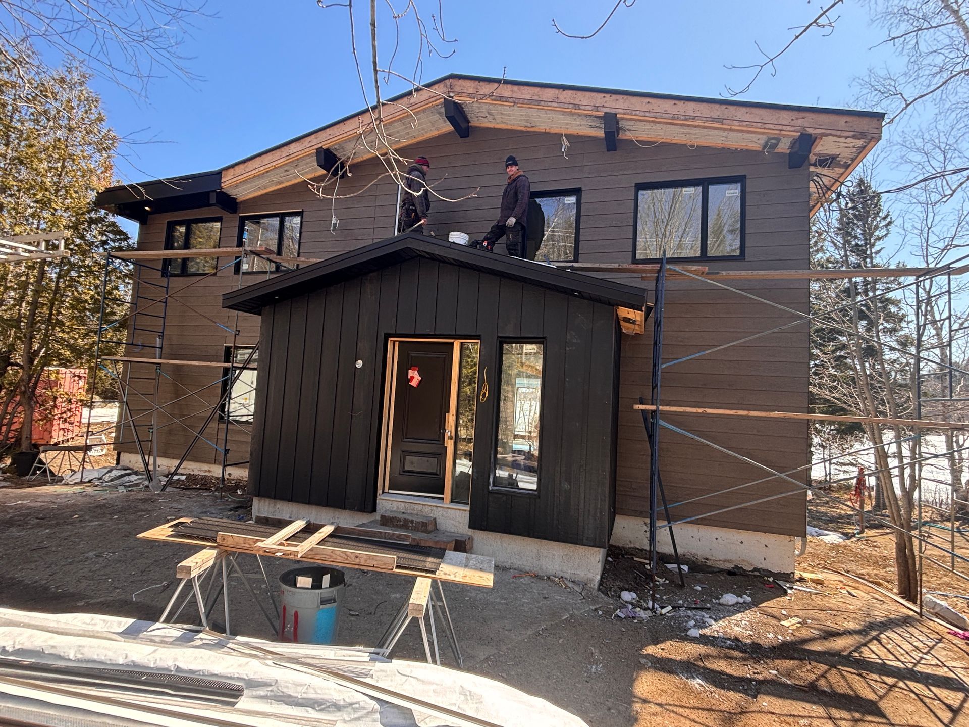Par une journée ensoleillée, des ouvriers, juchés sur des échafaudages, installent un bardage en bois foncé sur la façade d'une maison en construction.