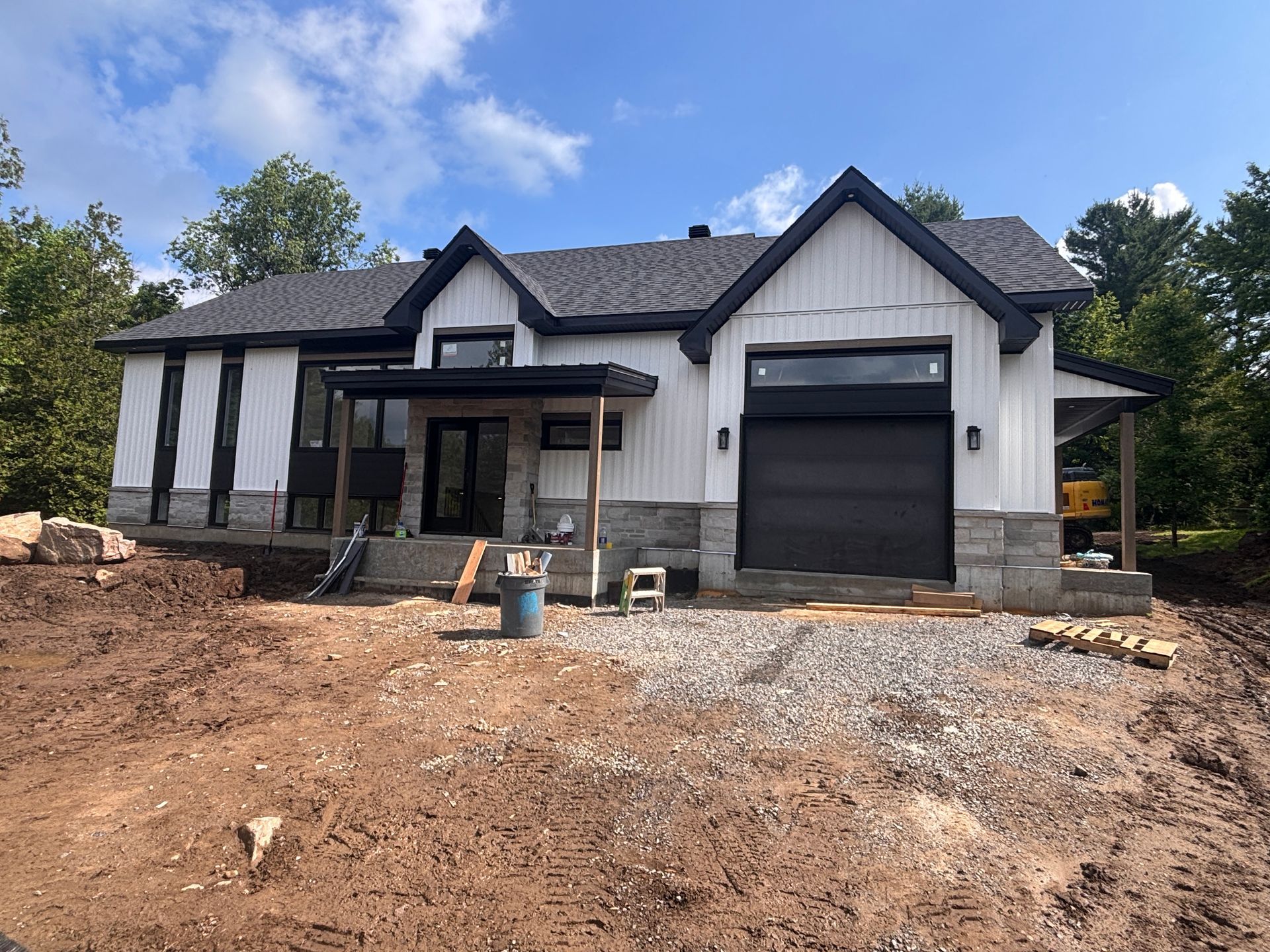Nouvelle maison de ferme moderne avec bardage vertical blanc, finitions noires et soubassement en pierre sous une allée partiellement aménagée.