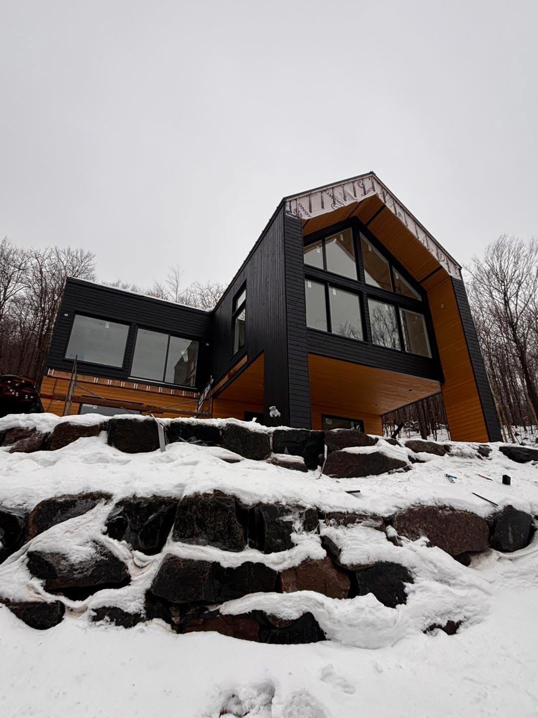 Une maison moderne, avec un bardage noir et des éléments en bois, se dresse sur un flanc de colline enneigé et rocailleux en terrasses, sous un ciel couvert.