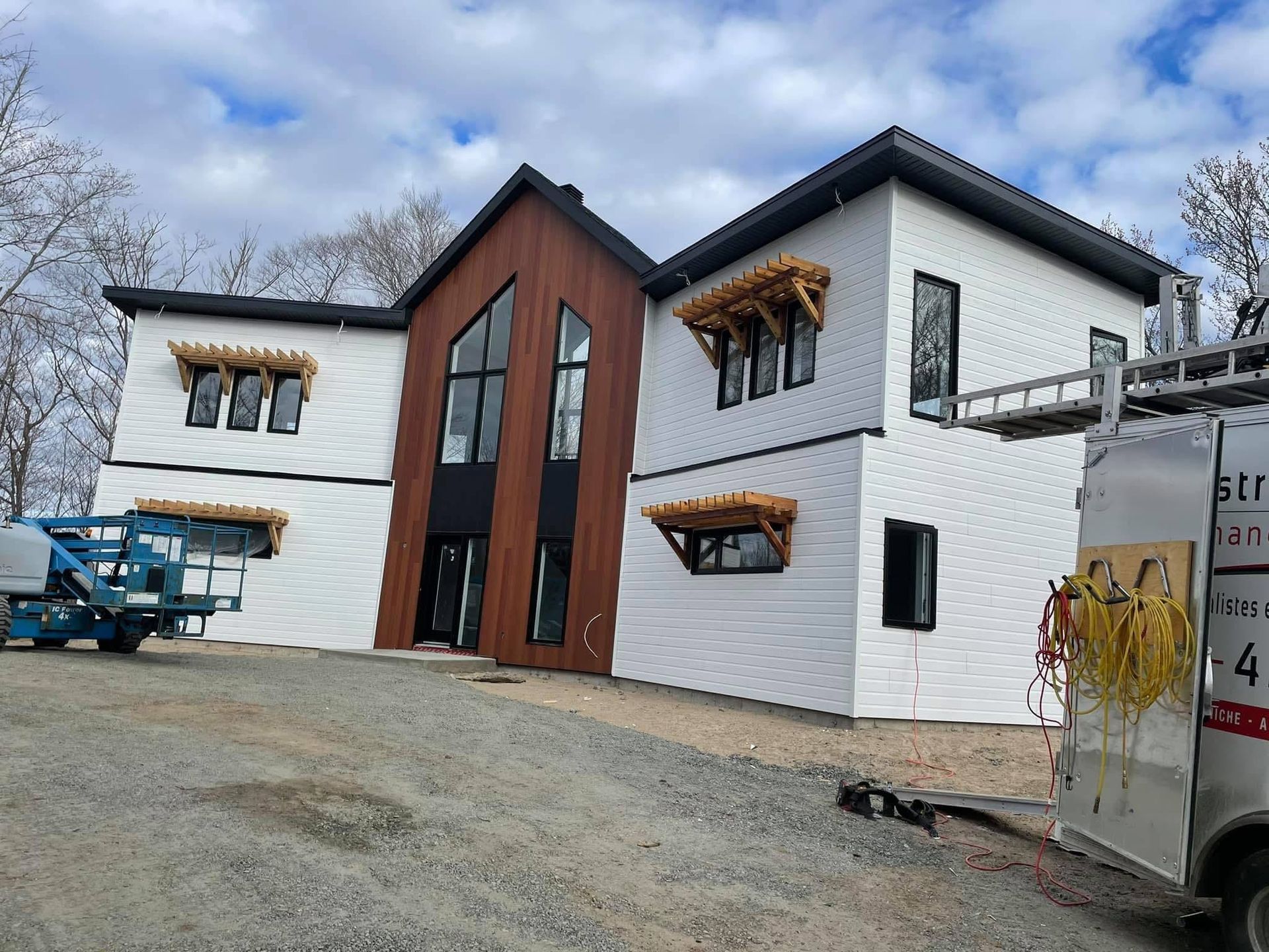 Une maison à deux étages en construction, avec un bardage blanc, un pignon central lambrissé et des auvents décoratifs en bois.